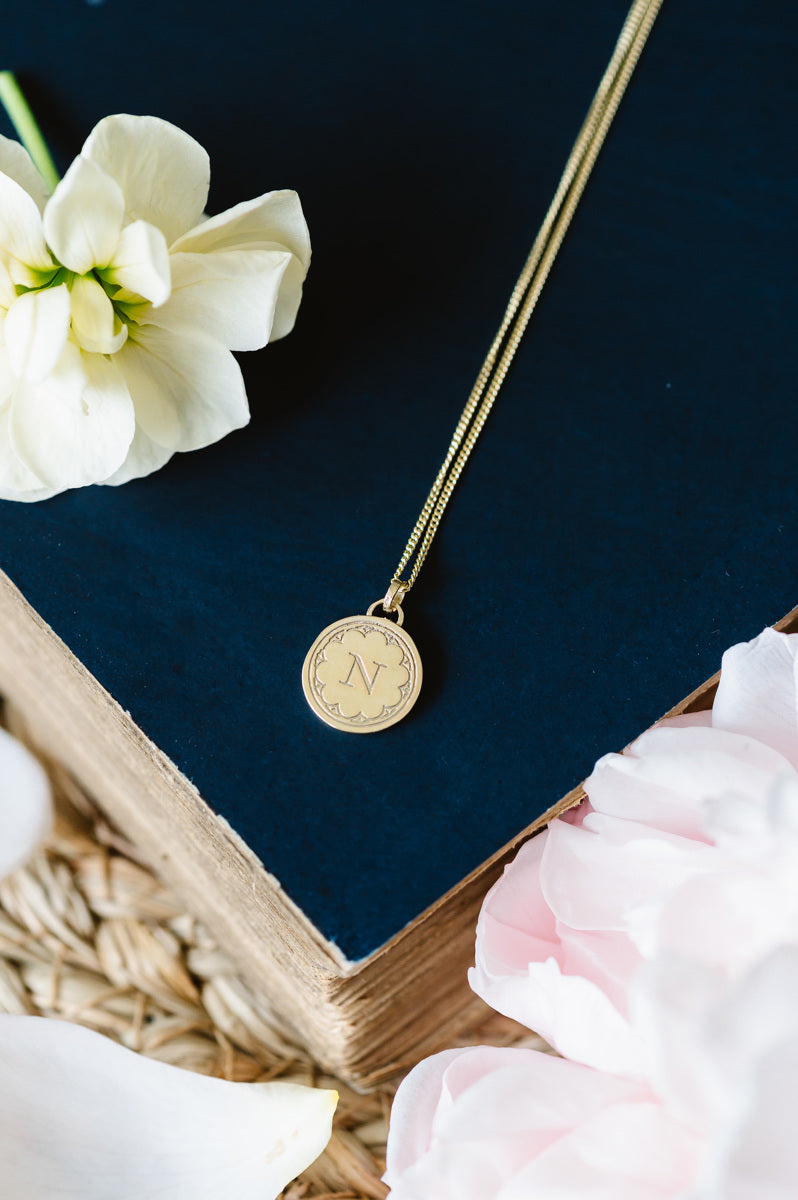 A dainty solid gold initial necklace sitting on a book, with some flowers in background.