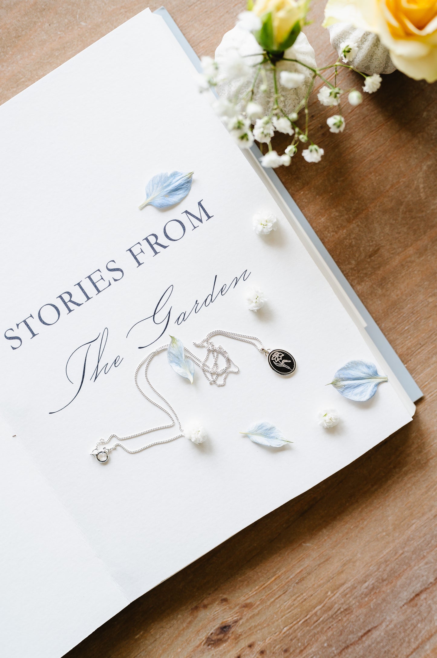 A dainty sterling silver necklace with a daffodil lying on an open book with scattered petals and flowers in the background.