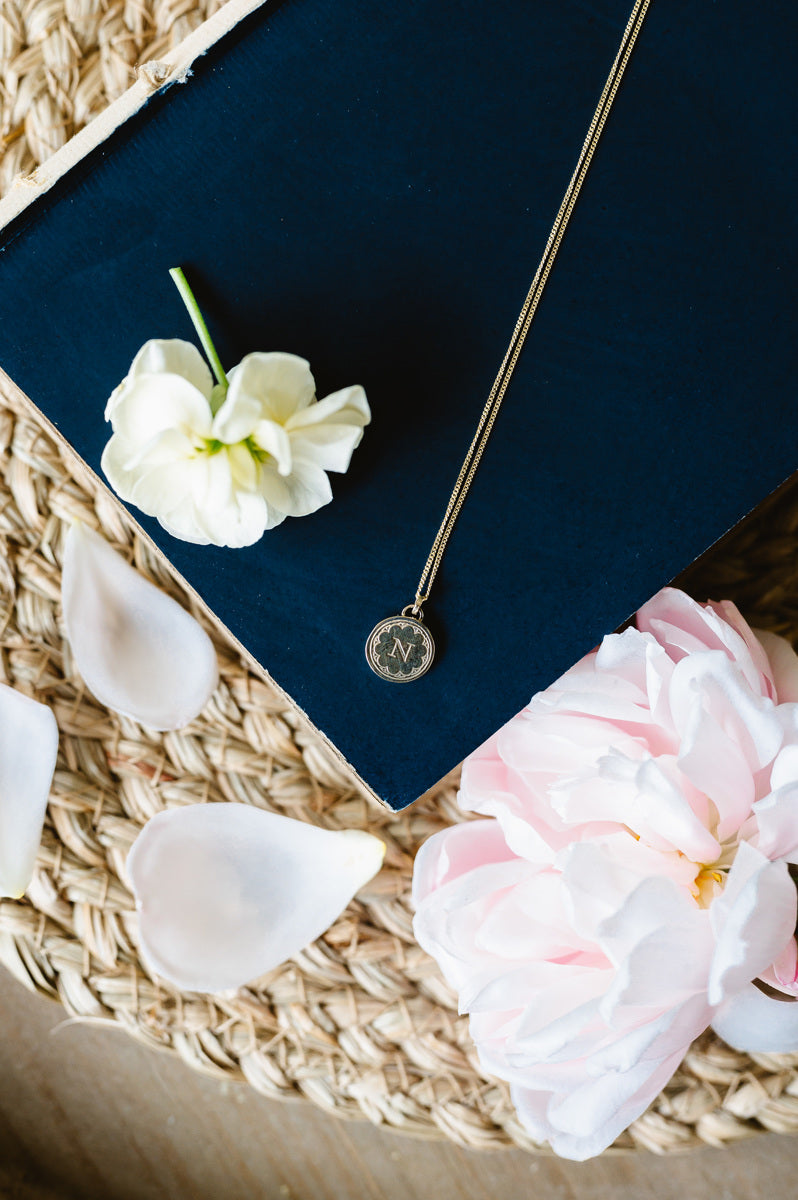 A dainty gold initial necklace sitting on a book with flowers and petals