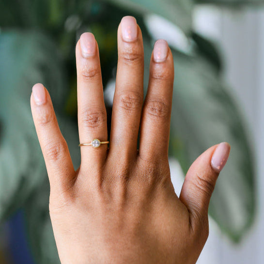 Diamond solitaire ring being worn, hand in front of a blurred green background.