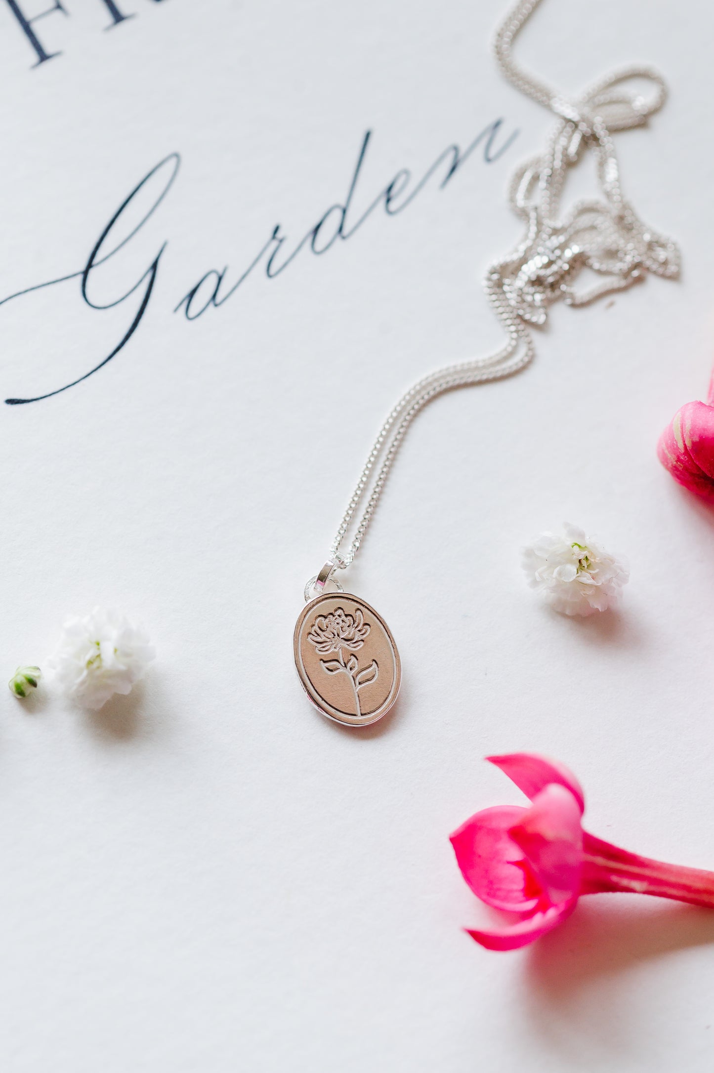 A sterling silver necklace with an oval coin pendant featuring a Chrysanthemum flower design. The dainty necklace is lying on a book with some flower buds.