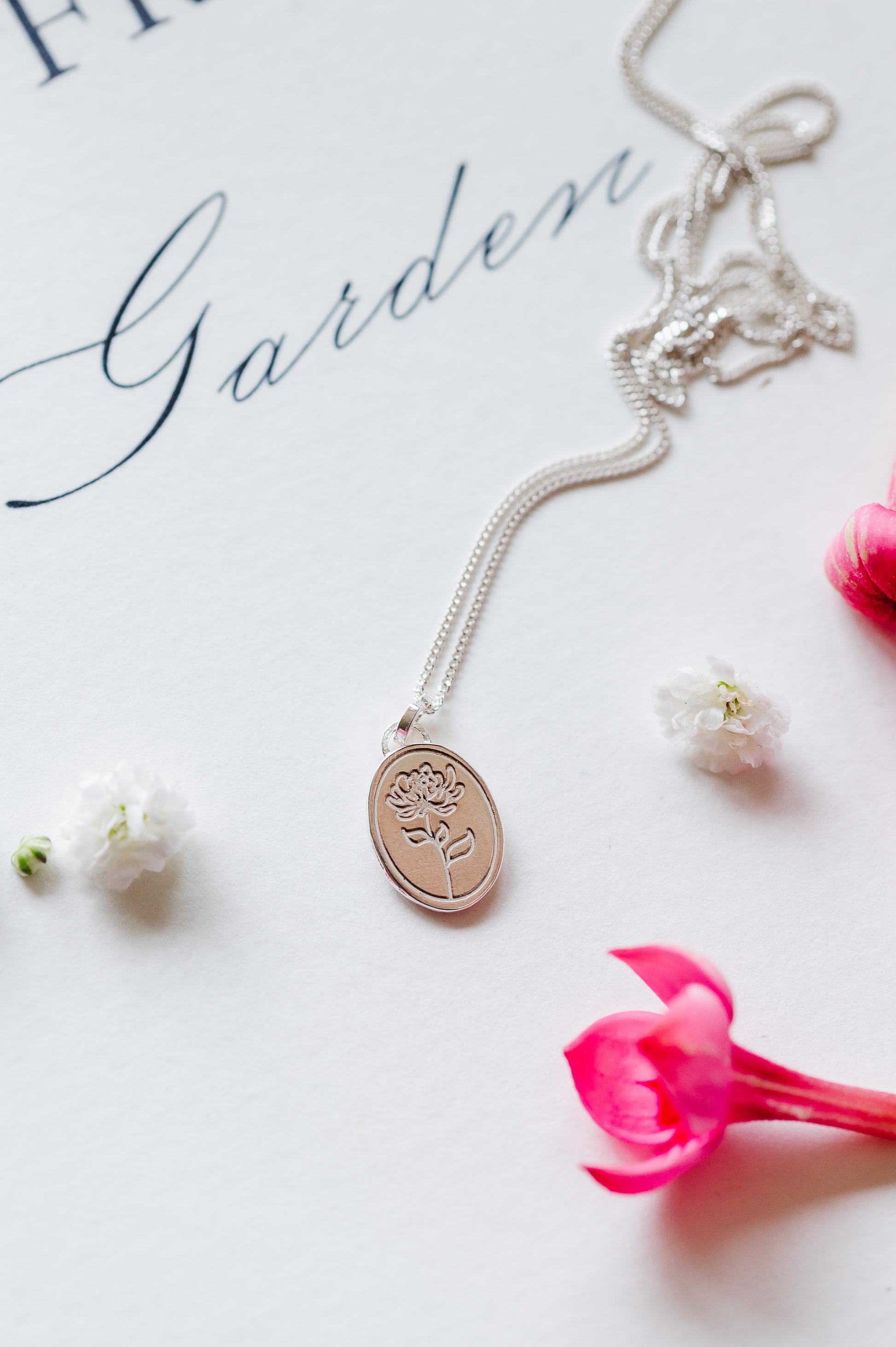 A sterling silver necklace with an oval coin pendant featuring a Chrysanthemum flower design. The dainty necklace is lying on a book with some flower buds.