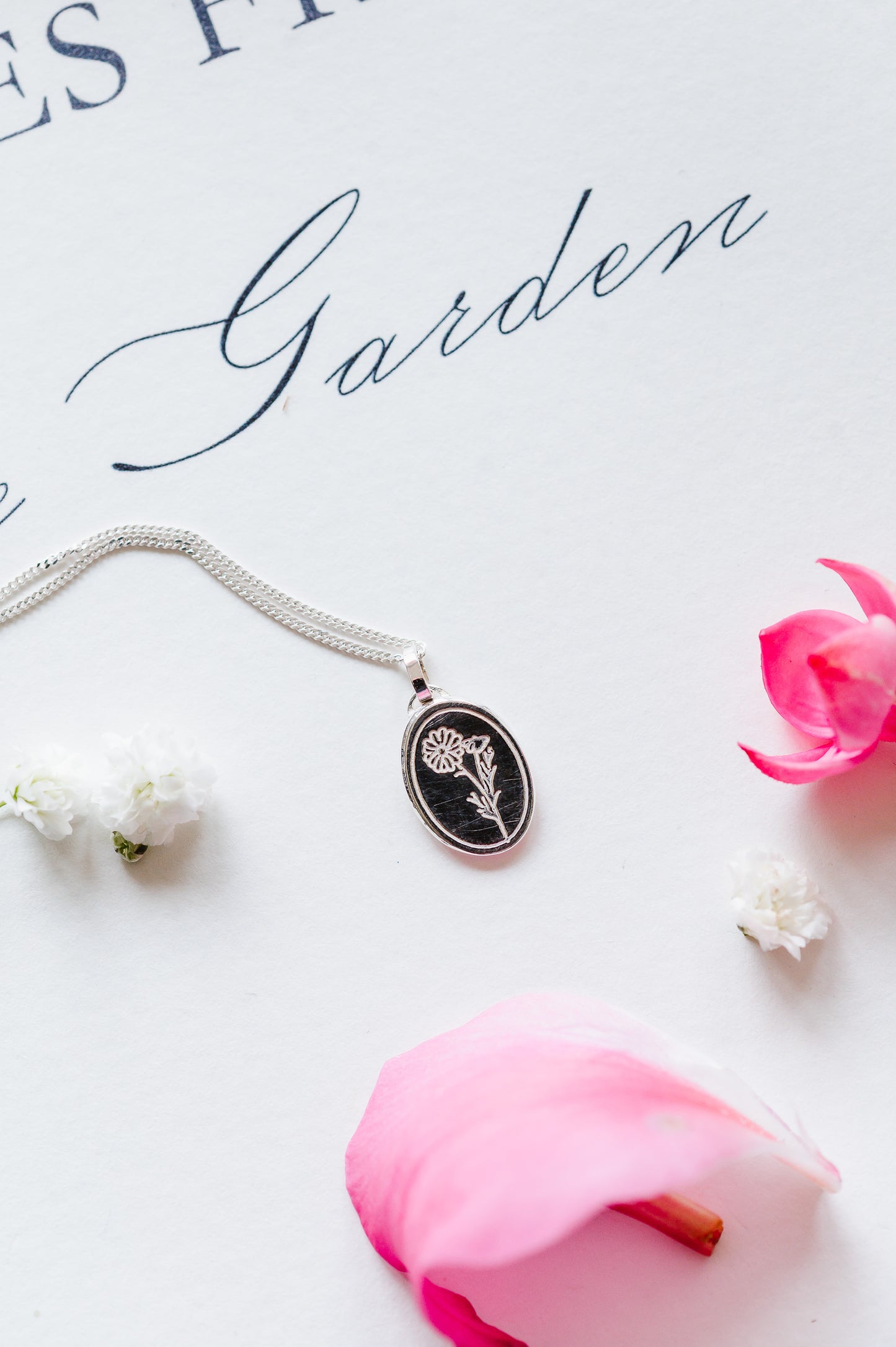 An oval coin necklace with a Cosmos flower design, displayed alongside a pink rose and a white flower, on a background with the text 'Garden'.