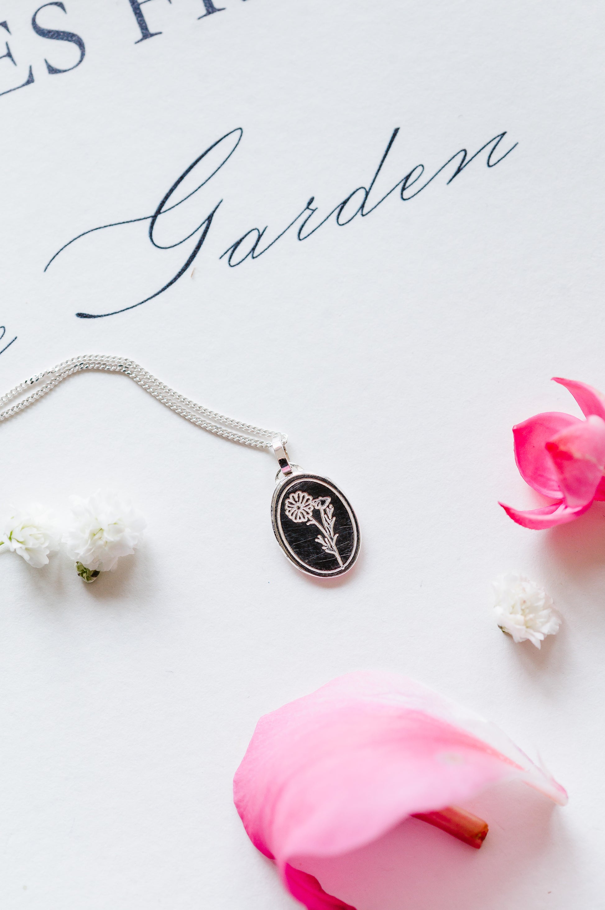 An oval coin necklace with a Cosmos flower design, displayed alongside a pink rose and a white flower, on a background with the text 'Garden'.