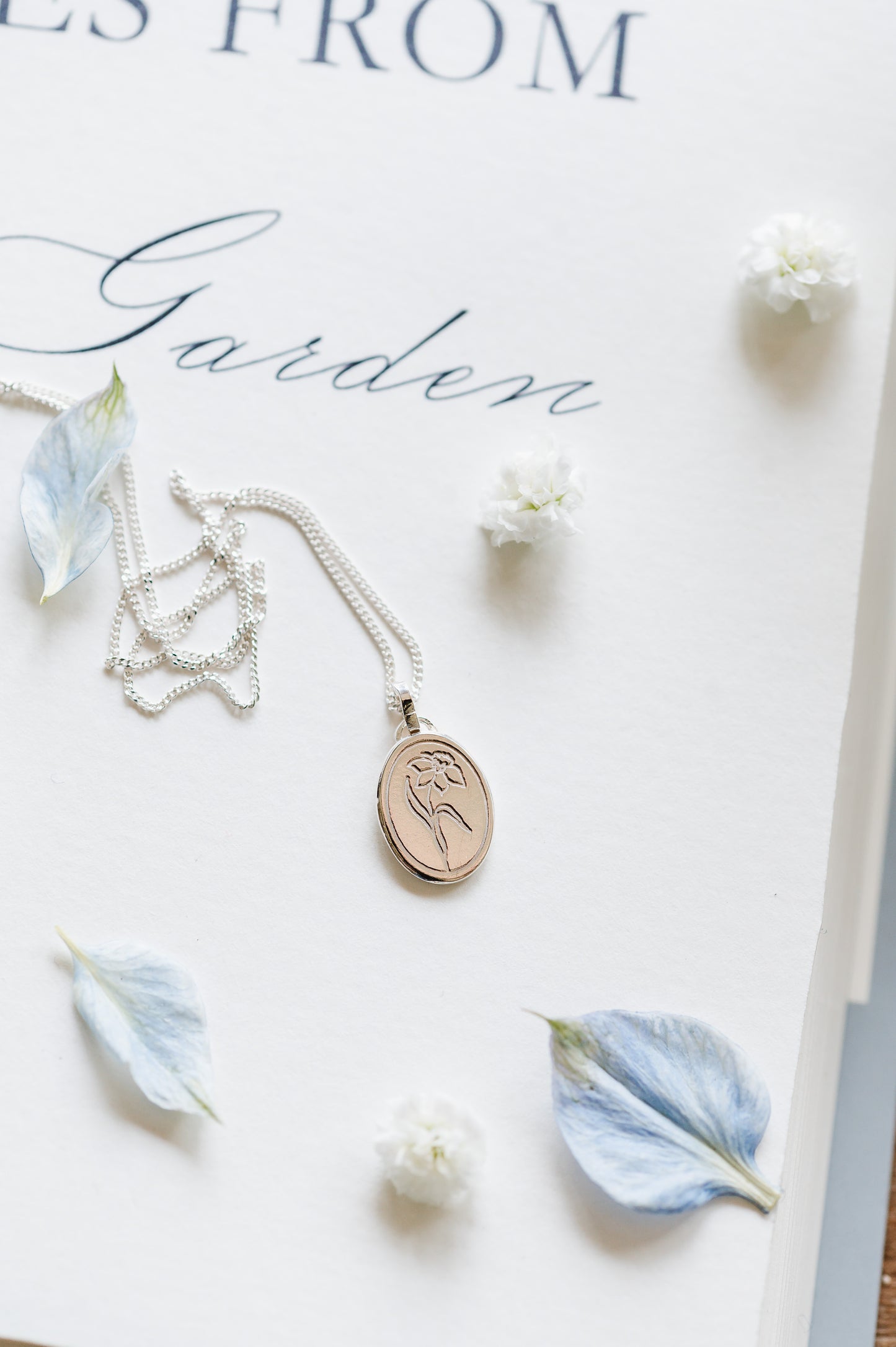 A sterling silver oval coin necklace with a daffodil design, displayed on a notebook with flower accents.