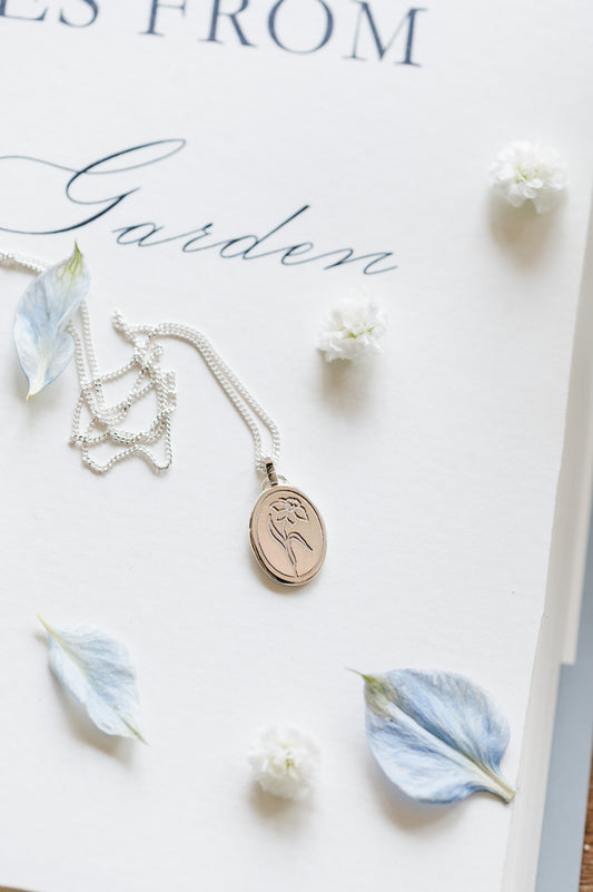 A sterling silver oval coin necklace with a daffodil design, displayed on a notebook with flower accents.