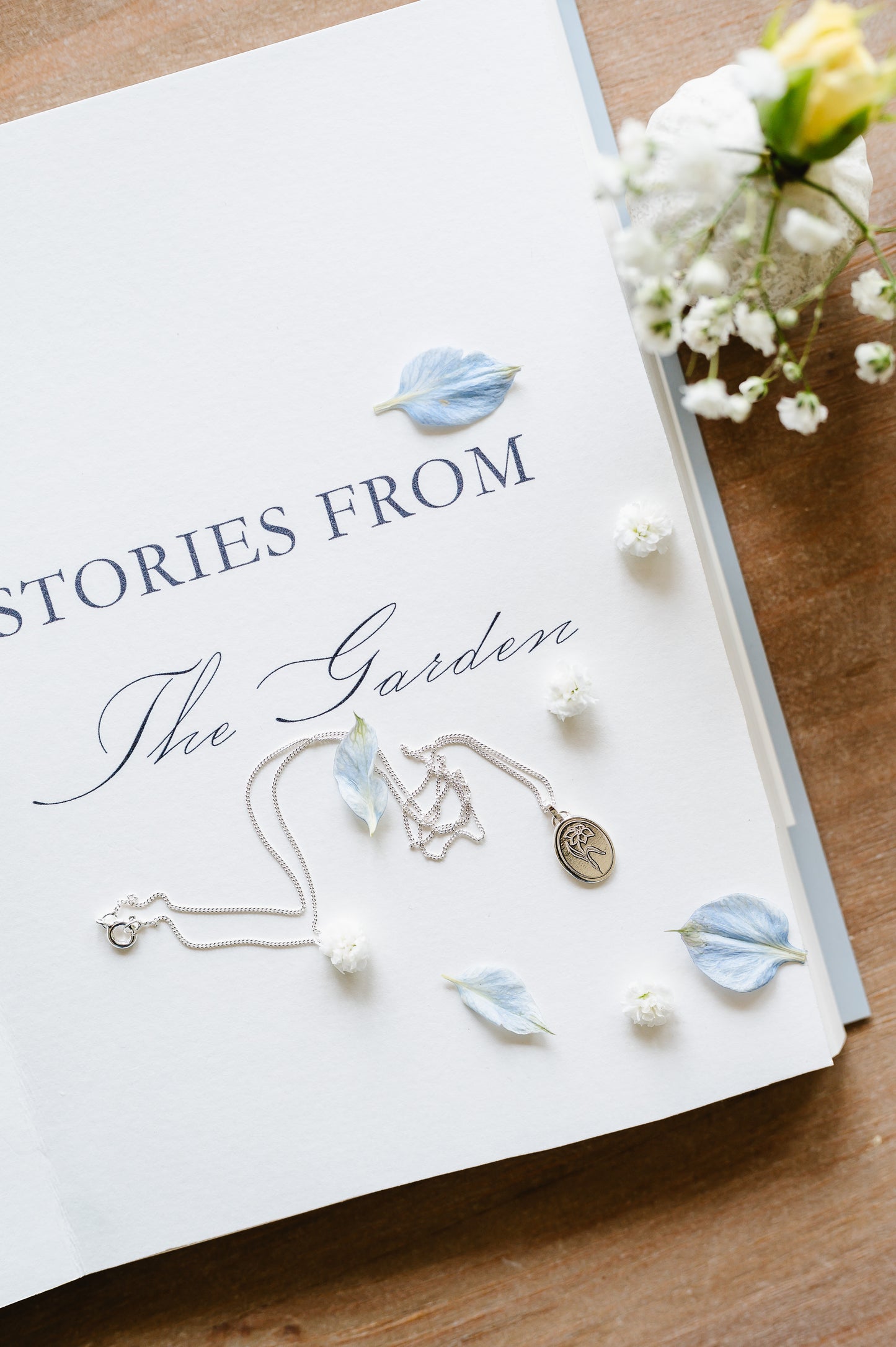 A dainty necklace with a daffodil in sterling silver, lying on an open book with scattered petals.