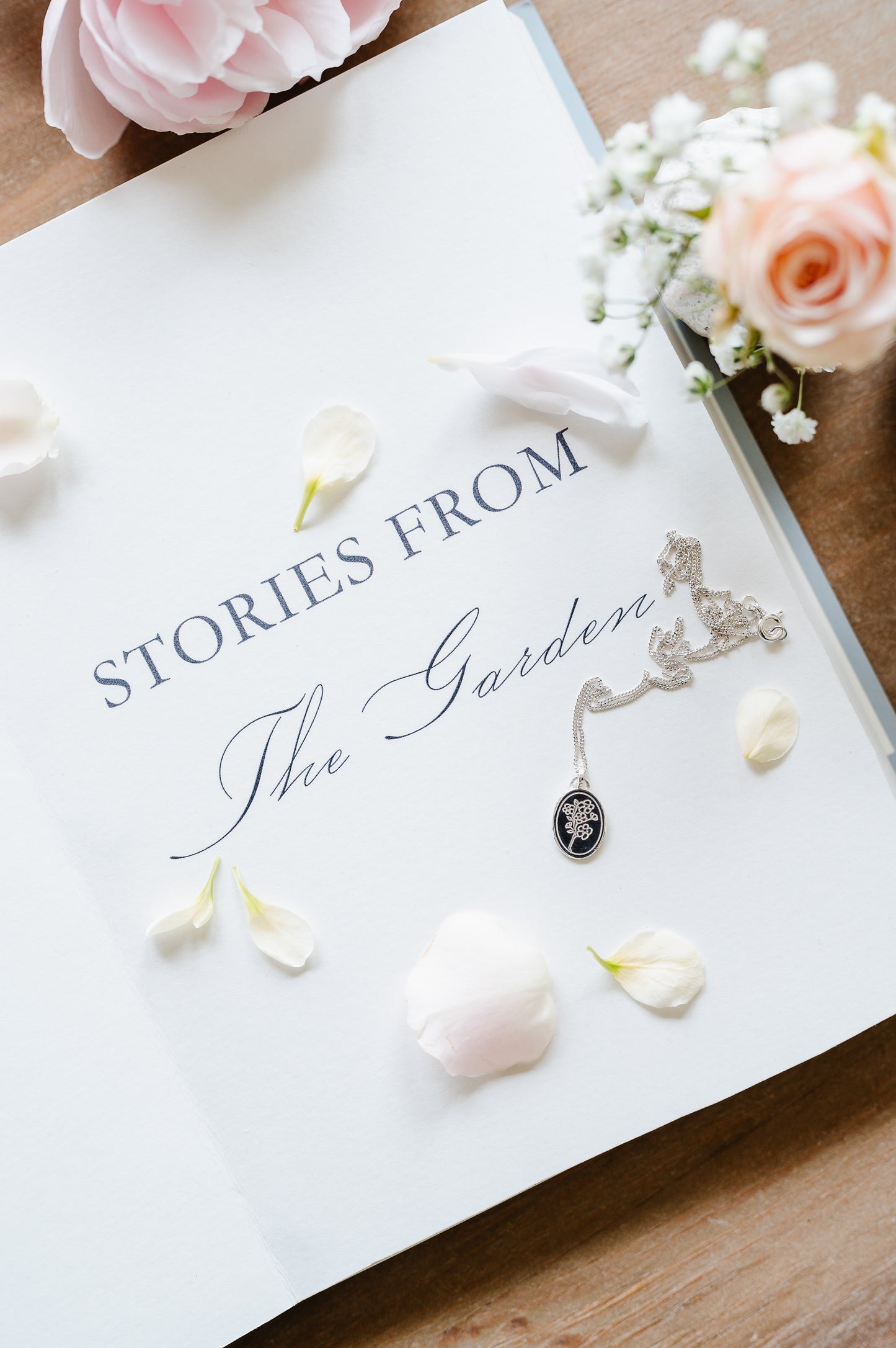 A dainty necklace in sterling silver with a cherry blossom design. The sterling silver necklace is lying on an open book with some flowers in the background.