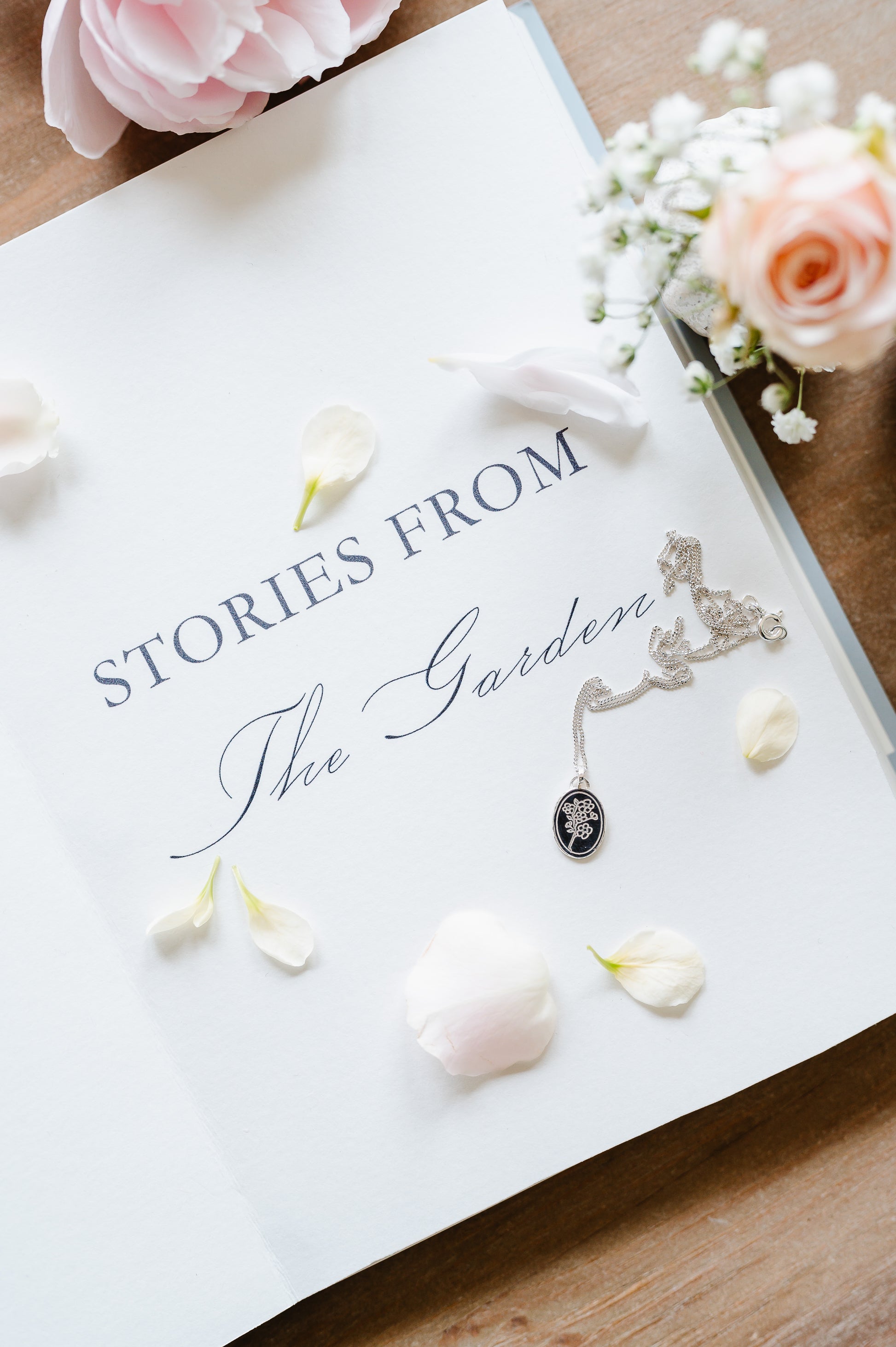 A dainty necklace in sterling silver with a cherry blossom design. The sterling silver necklace is lying on an open book with some flowers in the background.