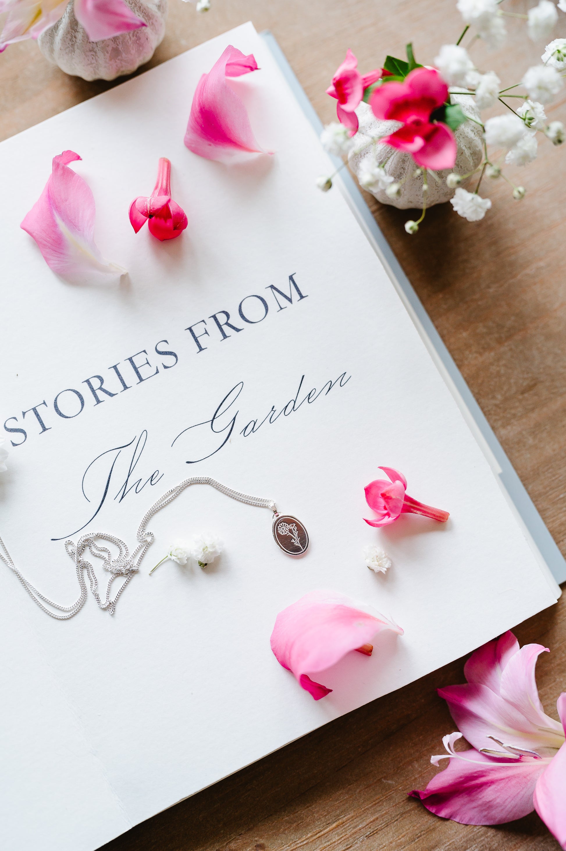 A dainty flower necklace in sterling silver with a cosmos, lying on an open book with some flowers in the background.