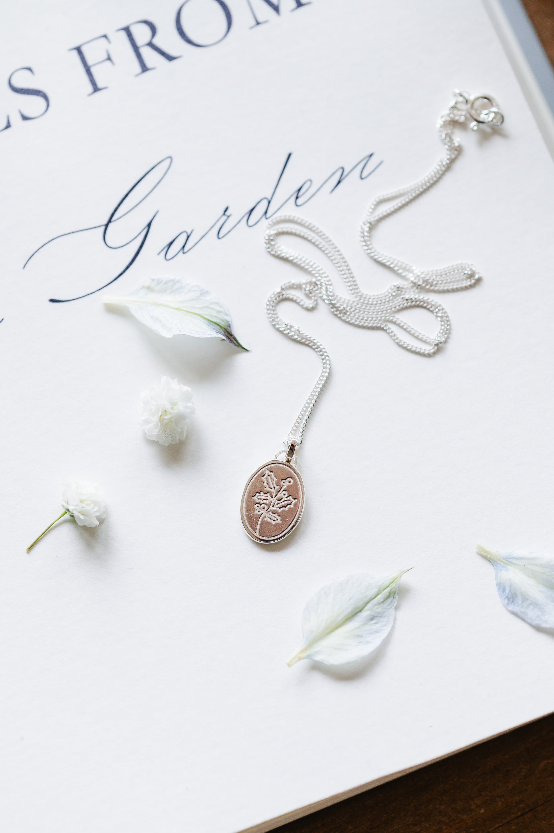 A dainty necklace with a holly sprig in sterling silver. Necklace is lying on a book with some loose petals.