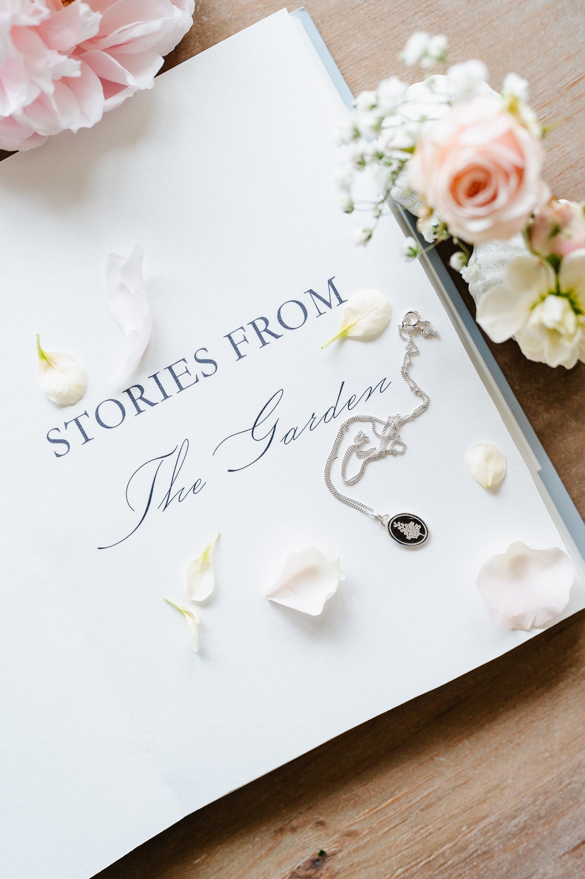 Delphinium necklace in sterling silver, lying on an open book with loose petals and flowers.