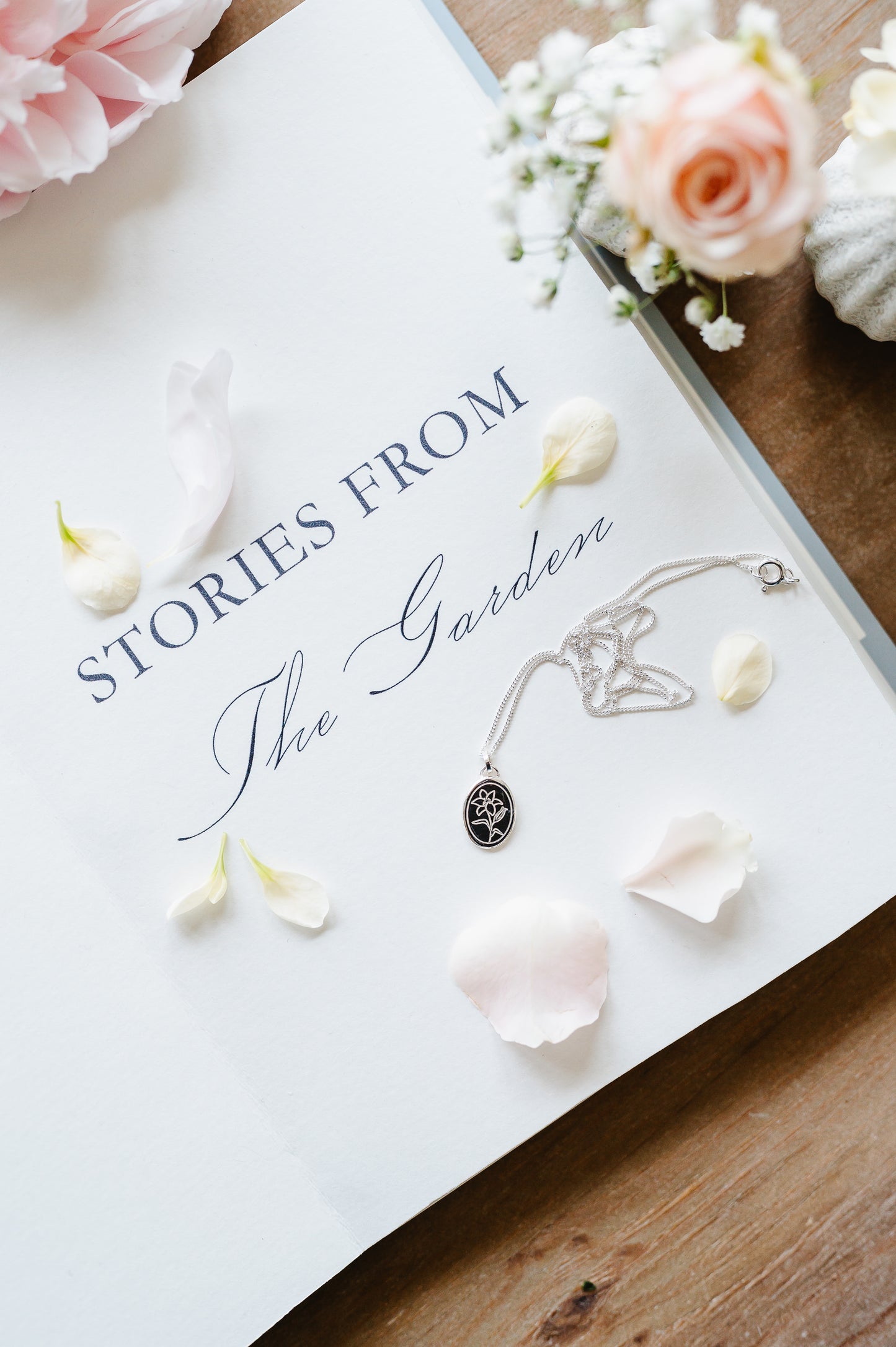 A delicate flower necklace with a lily in sterling silver, lying on an open book with flowers in the background.