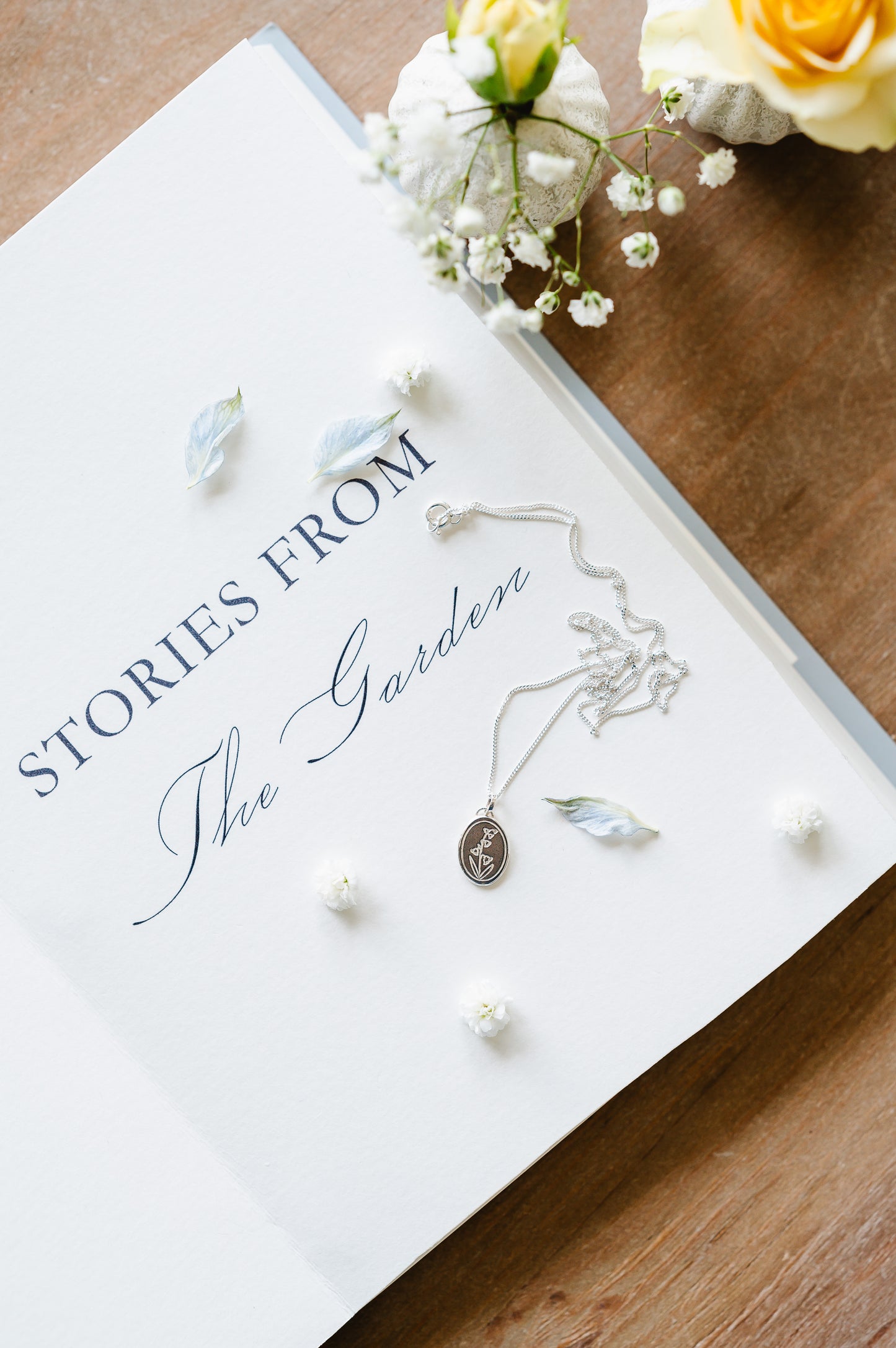 A dainty necklace in sterling silver with a lily of the valley design. The necklace is lying on a book, with flowers in the background. 