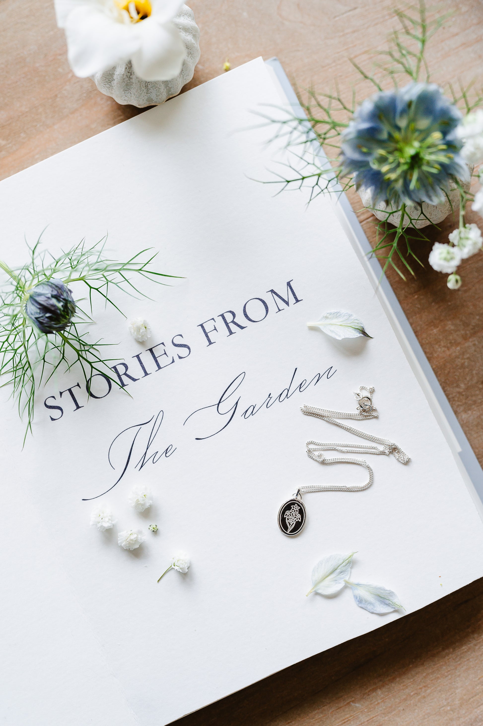 A dainty flower necklace with a bunch of narcissus in sterling silver. The necklace is lying on a book page with some flowers.