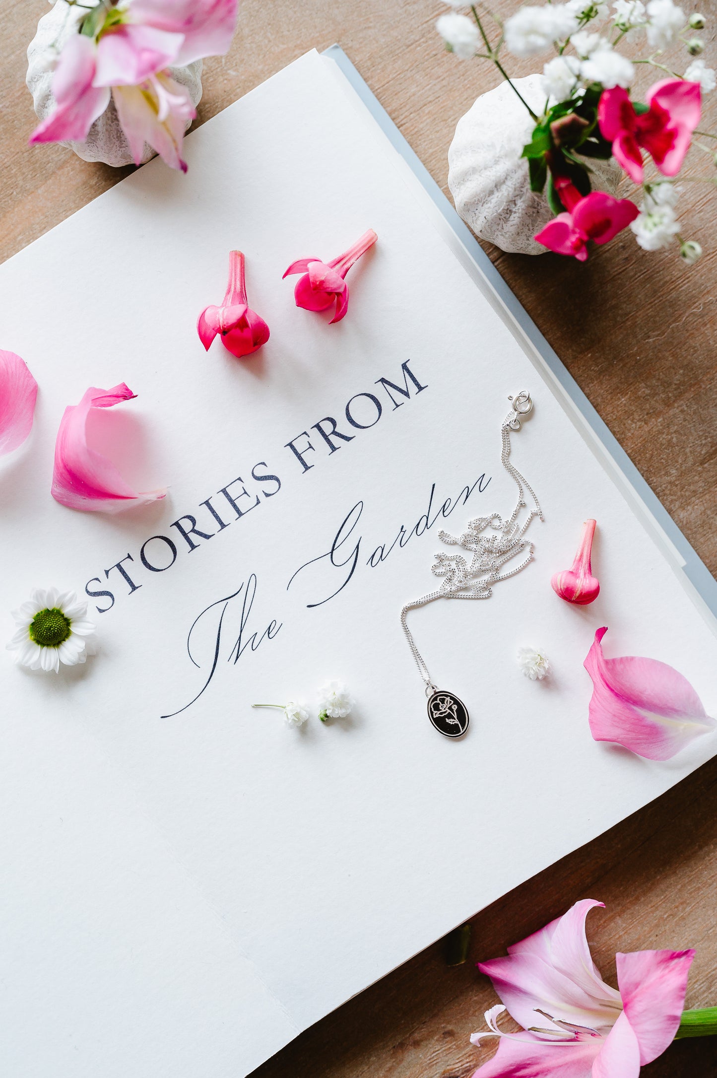 A dainty flower necklace with a poppy in sterling silver. The necklace is lying on an open book with flowers in the background. 
