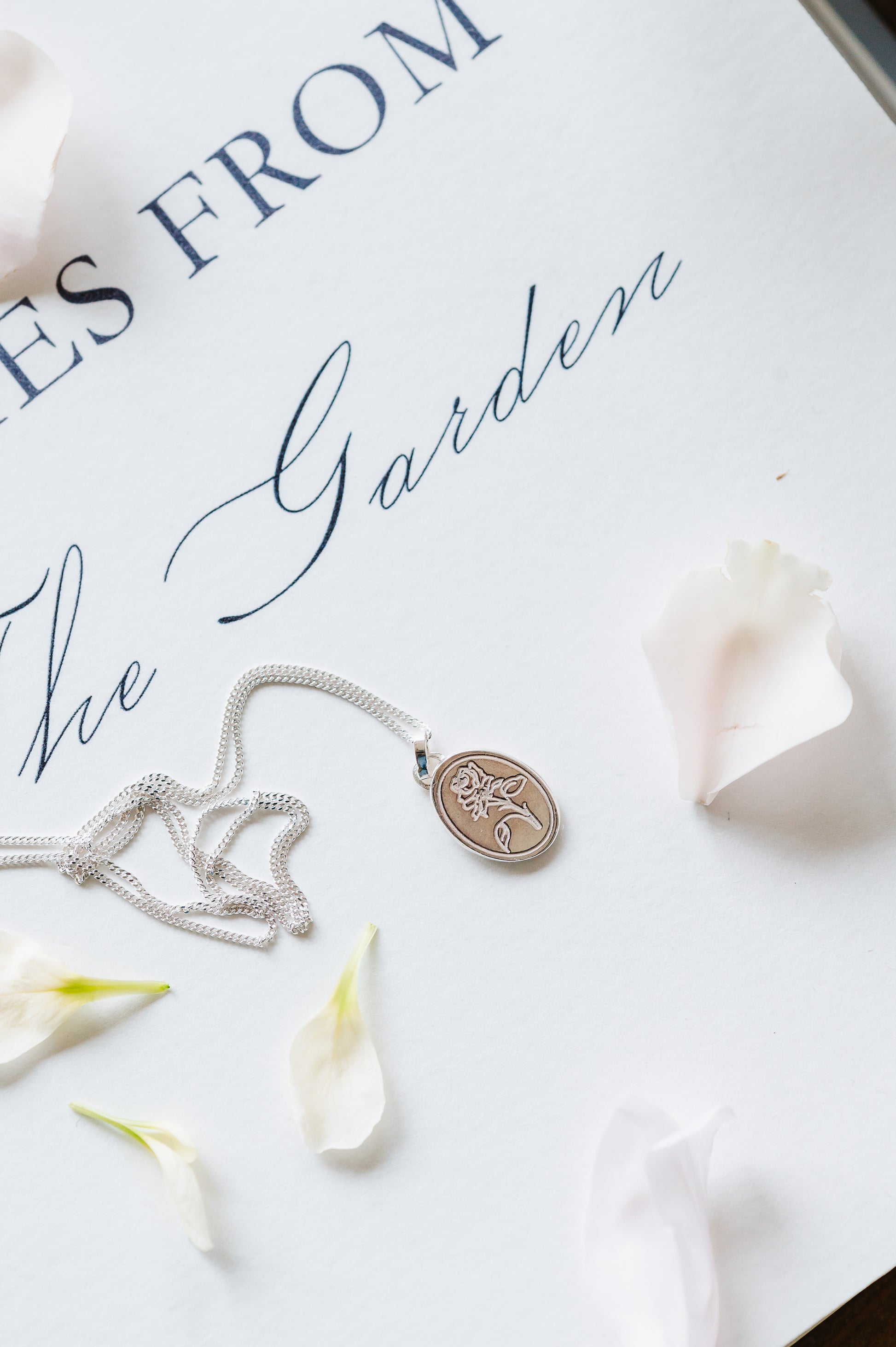 A dainty flower necklace with a rose in sterling silver, lying on a white background with some petals.