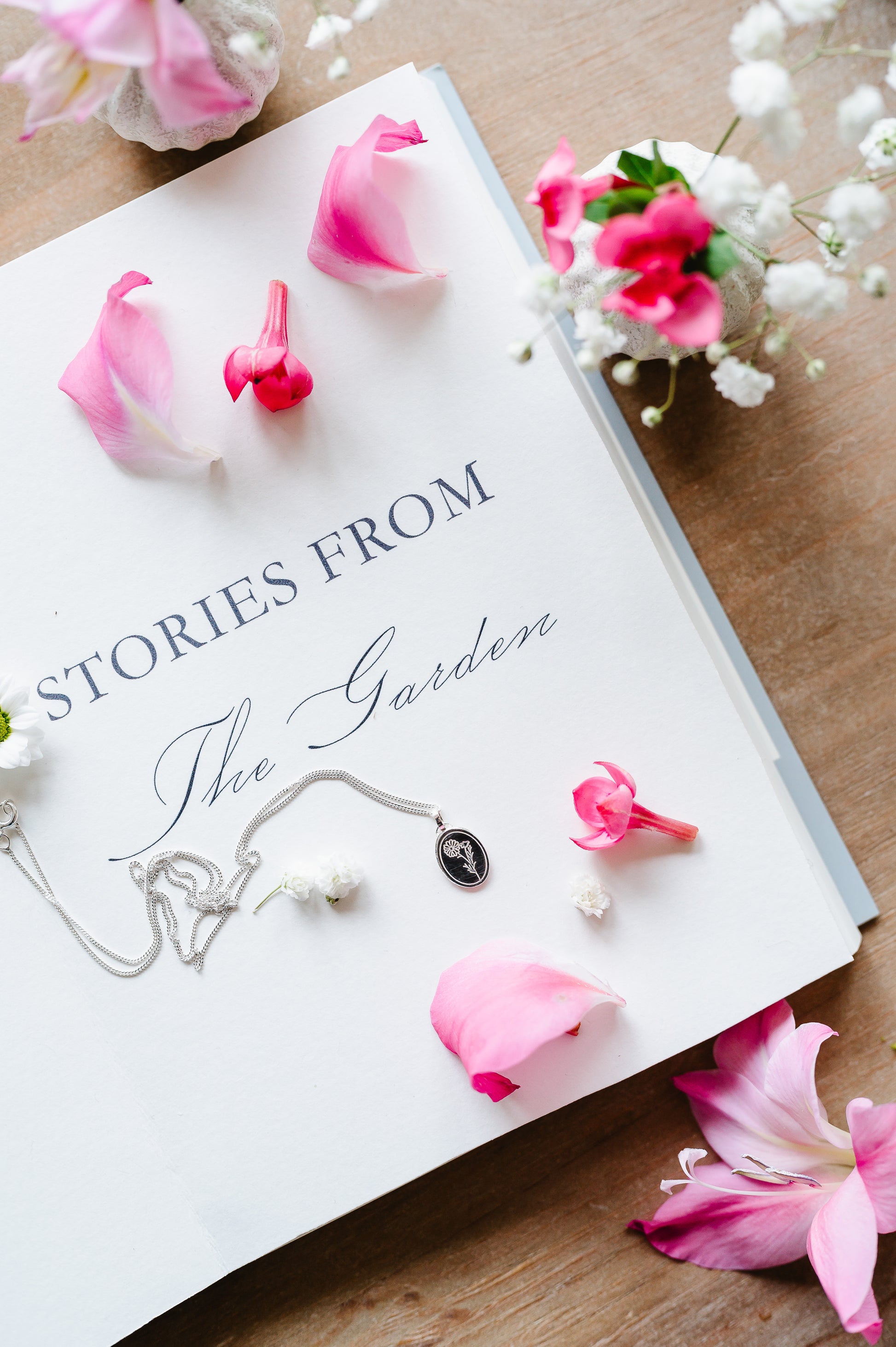 A sterling silver necklace with a cosmos flower, lying on an open book with some flowers