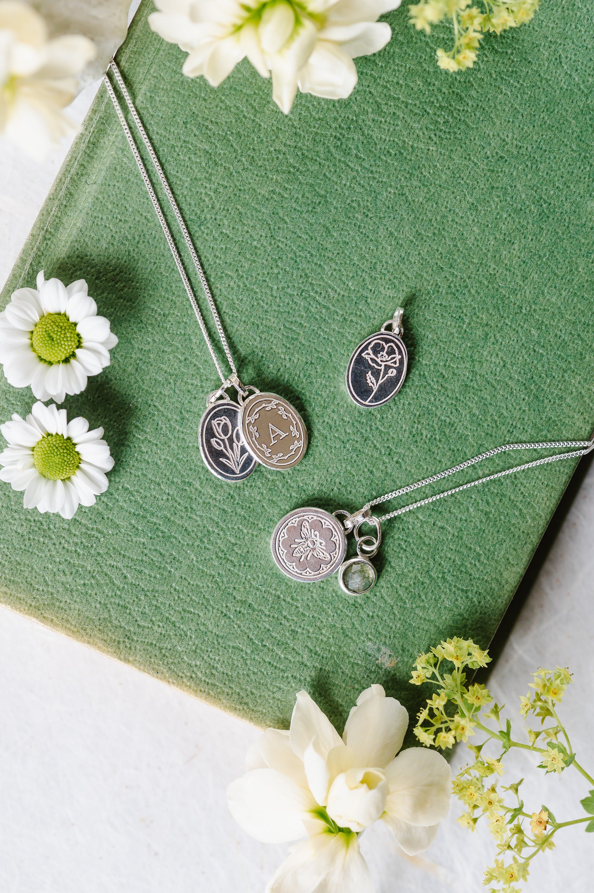A selection of sterling silver necklaces lying on a book cover. Engraved flower necklaces, an initial necklace and a teeny bee.