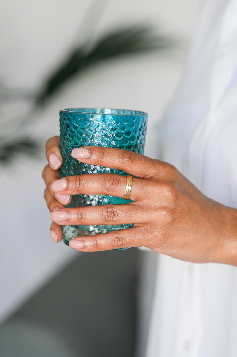 A hand holding a glass, wearing a gold wedding ring with grooved details.