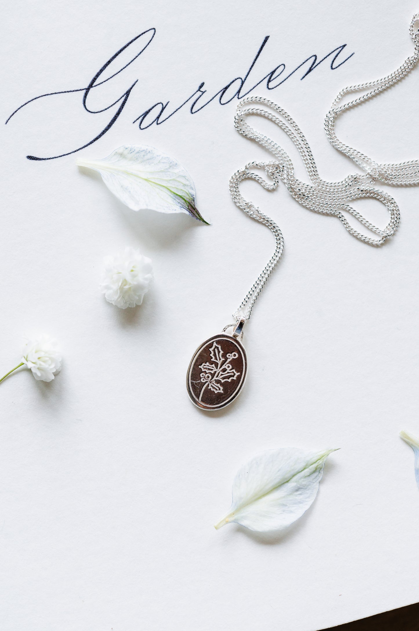 A sterling silver necklace with a holly sprig, the dainty necklace is lying on a white background with loose petals. 