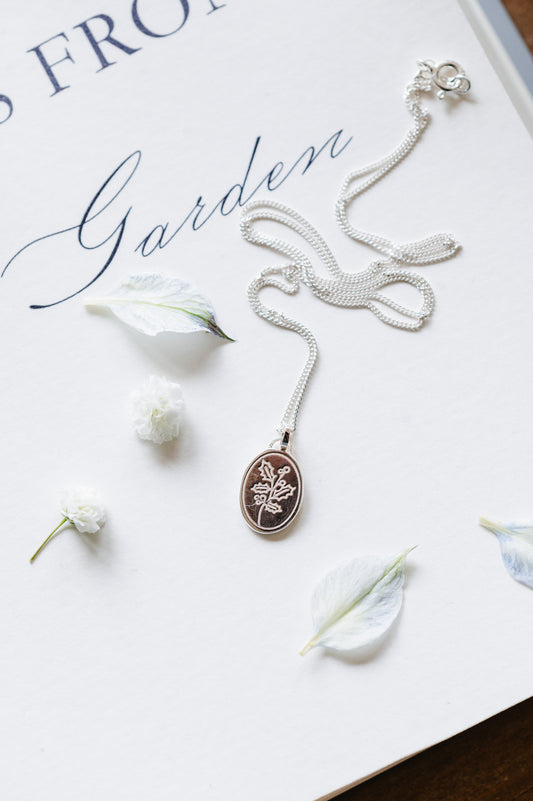 A sterling silver necklace with an oval pendant featuring a Holly sprig design, displayed on a book with the text 'Garden'. 