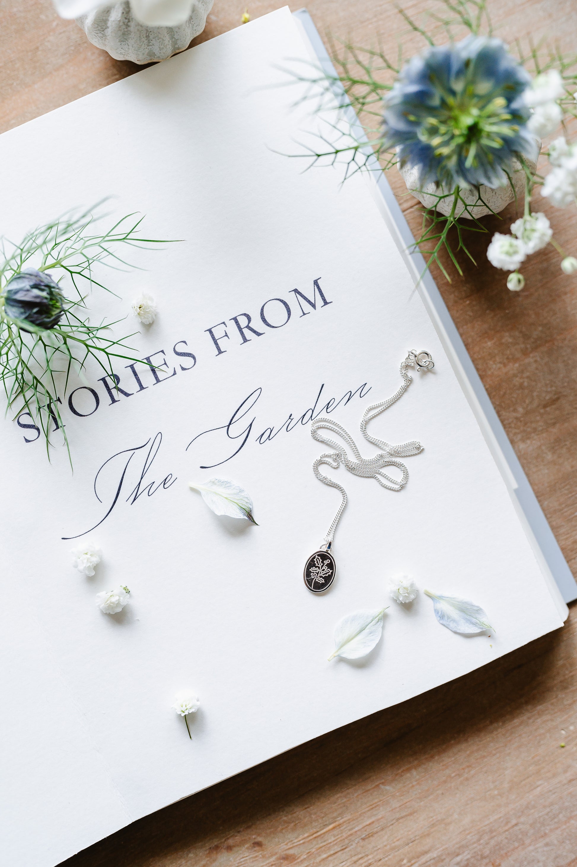 Dainty flower necklace with holly sprig in sterling silver. The necklace is lying on a book with flowers in the background.