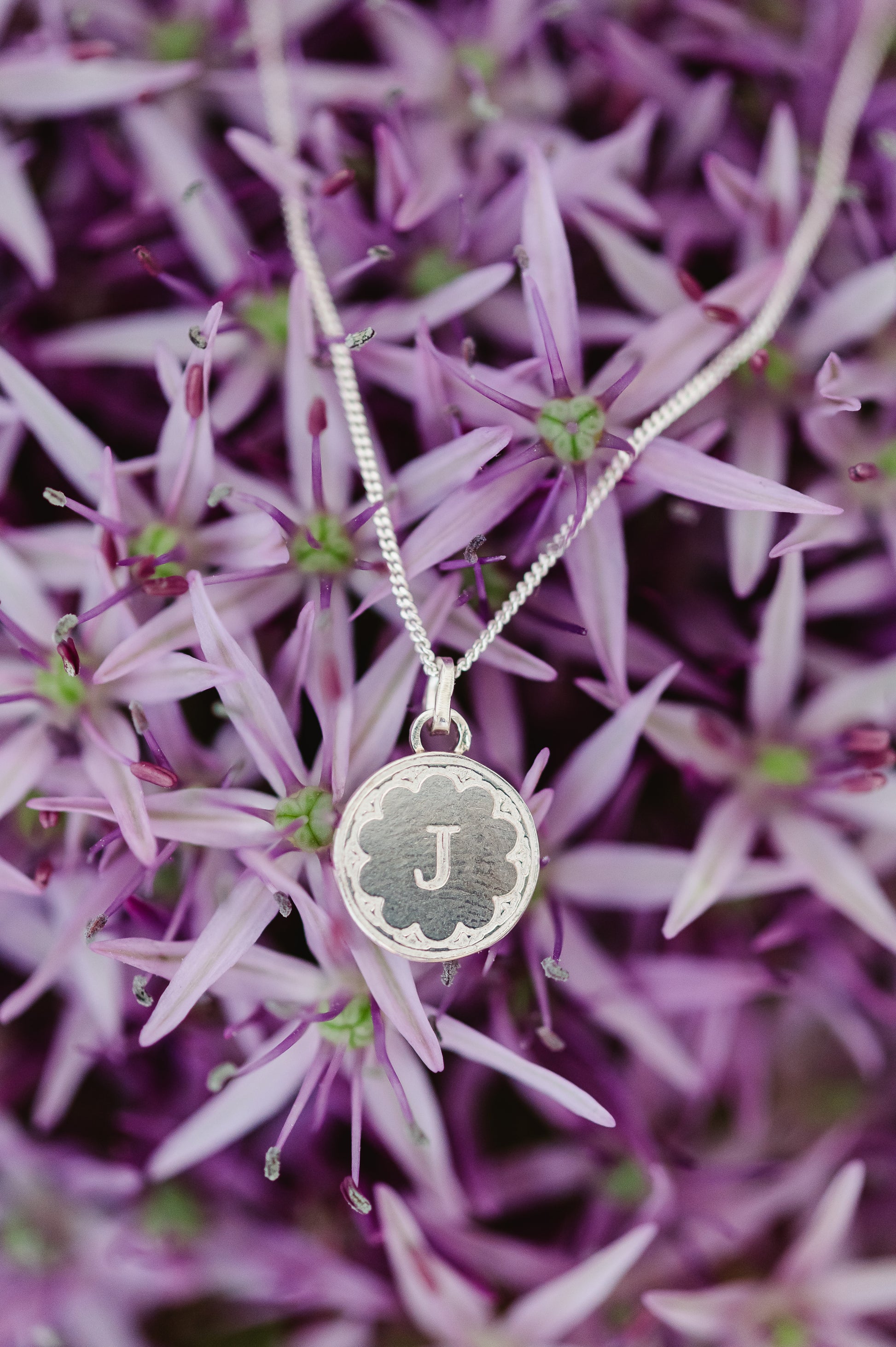 A round initial necklace in sterling silver, shown hanging on a very large allium flower