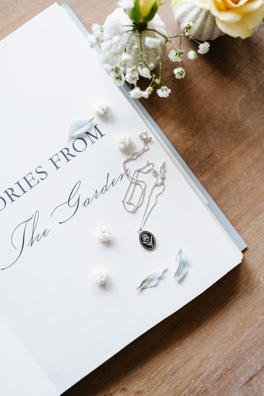 A dainty flower necklace with an iris in sterling silver, lying on a book with flowers in the background.
