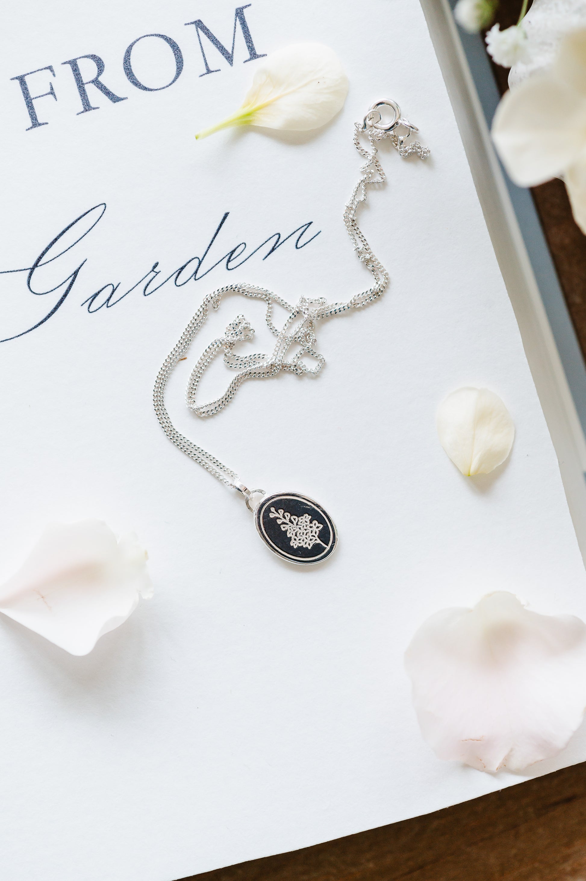 A sterling silver necklace with an oval coin pendant featuring a Larkspur/Delphinium design, displayed on a white surface with petals around it.