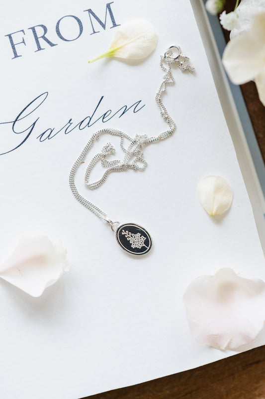 A sterling silver necklace with an oval coin pendant featuring a Larkspur/Delphinium design, displayed on a white surface with petals around it.