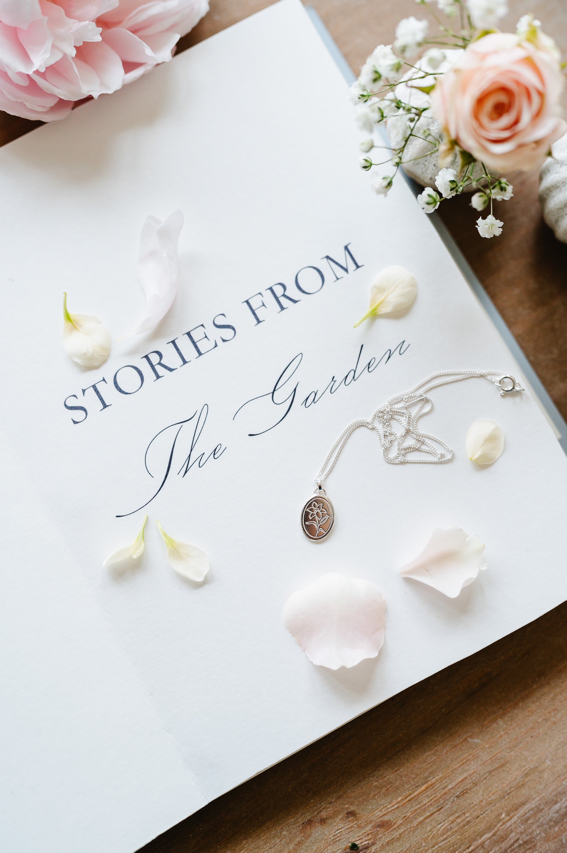 A sterling silver necklace with an oval coin design featuring a lily flower, displayed on a book with flowers.