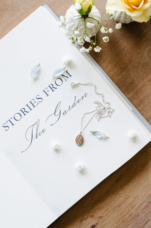 A sterling silver necklace featuring an oval coin with a Lily of the valley design, displayed on a book with a floral arrangement.