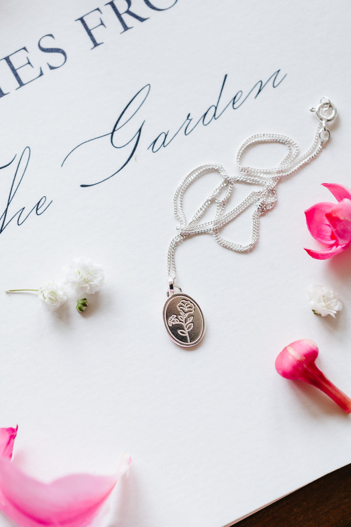 A sterling silver necklace with an oval coin pendant featuring a Morning Glory flower design, displayed on a white surface with a few pink flowers around it. 