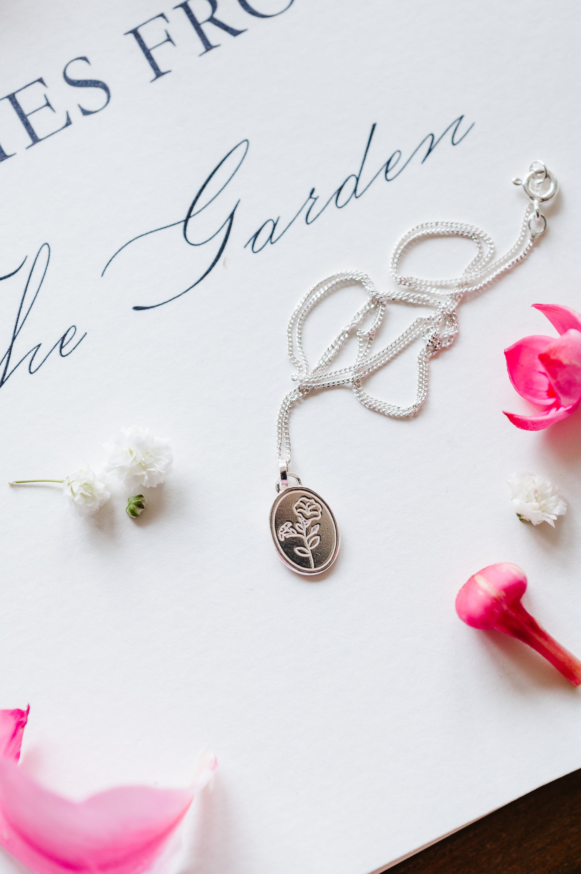A sterling silver necklace with an oval coin pendant featuring a Morning Glory flower design, displayed on a white surface with a few pink flowers around it. 