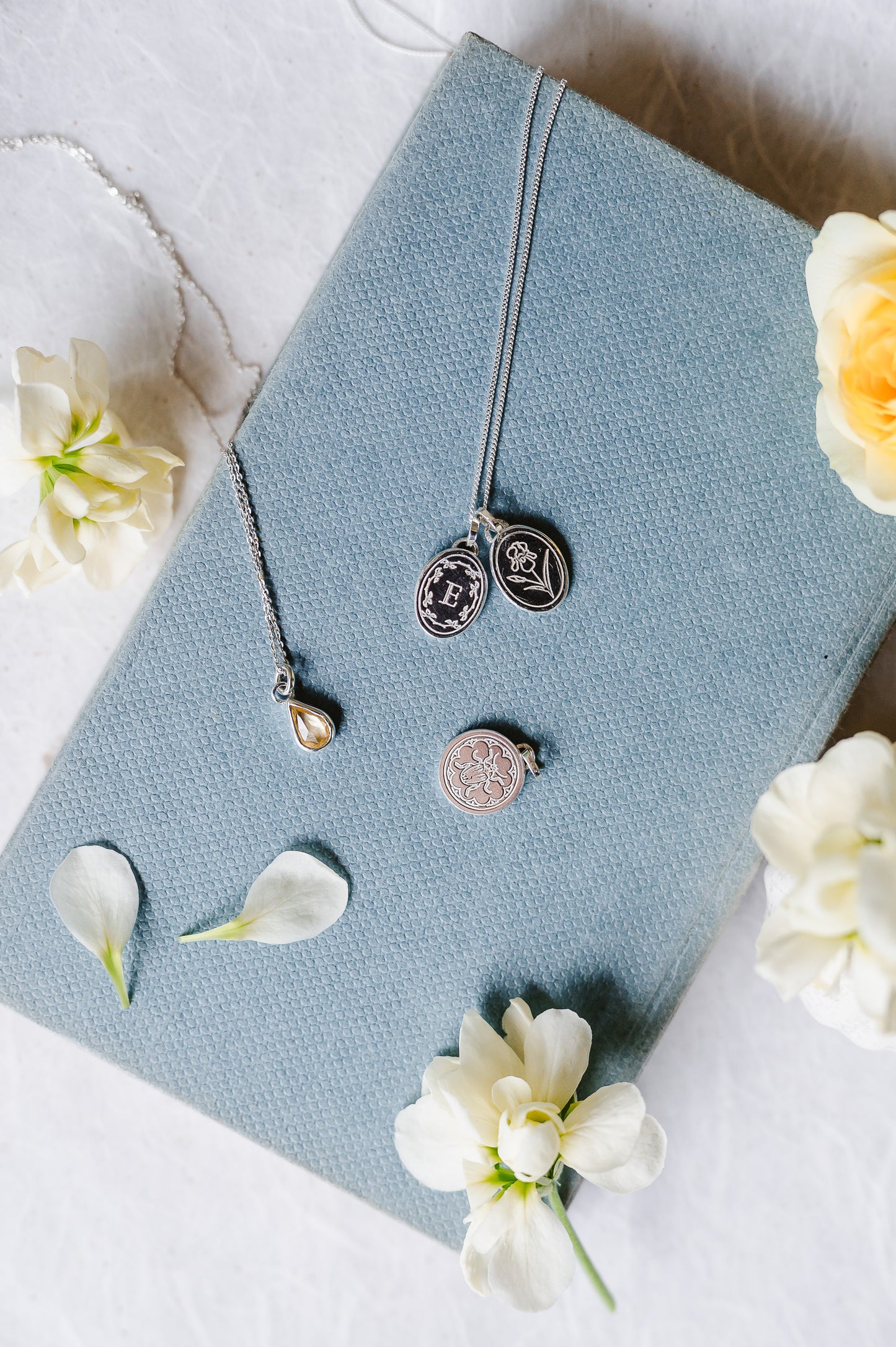 An oval initial necklace in sterling silver, with iris necklace on a chain. A beetle charm and a sapphire teardrop also shown, lying on a book cover with flowers.