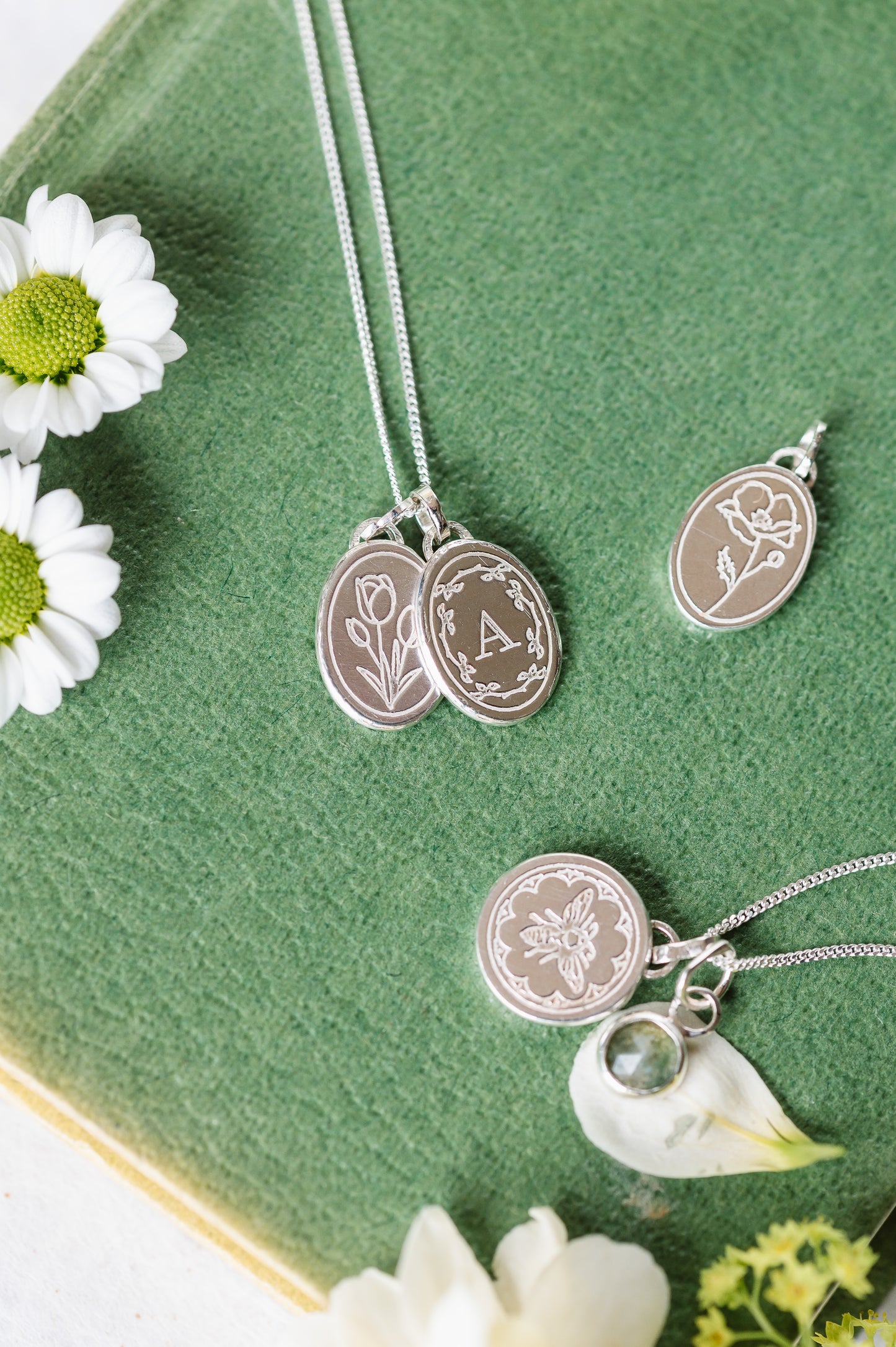 Oval initial necklace and tulip necklace, shown with some other engraved silver necklaces. All lying on a book with some flowers.