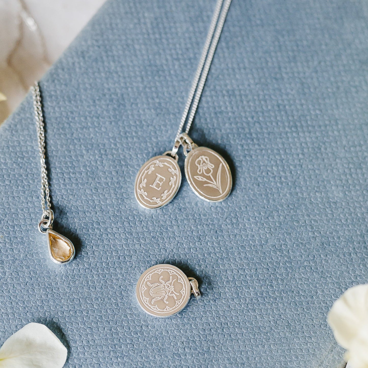 Sterling silver initial necklace stacked with Iris birth flower. A bee and a yellow teardrop charm also shown. All sterling silver pieces lying on a pale blue book cover.