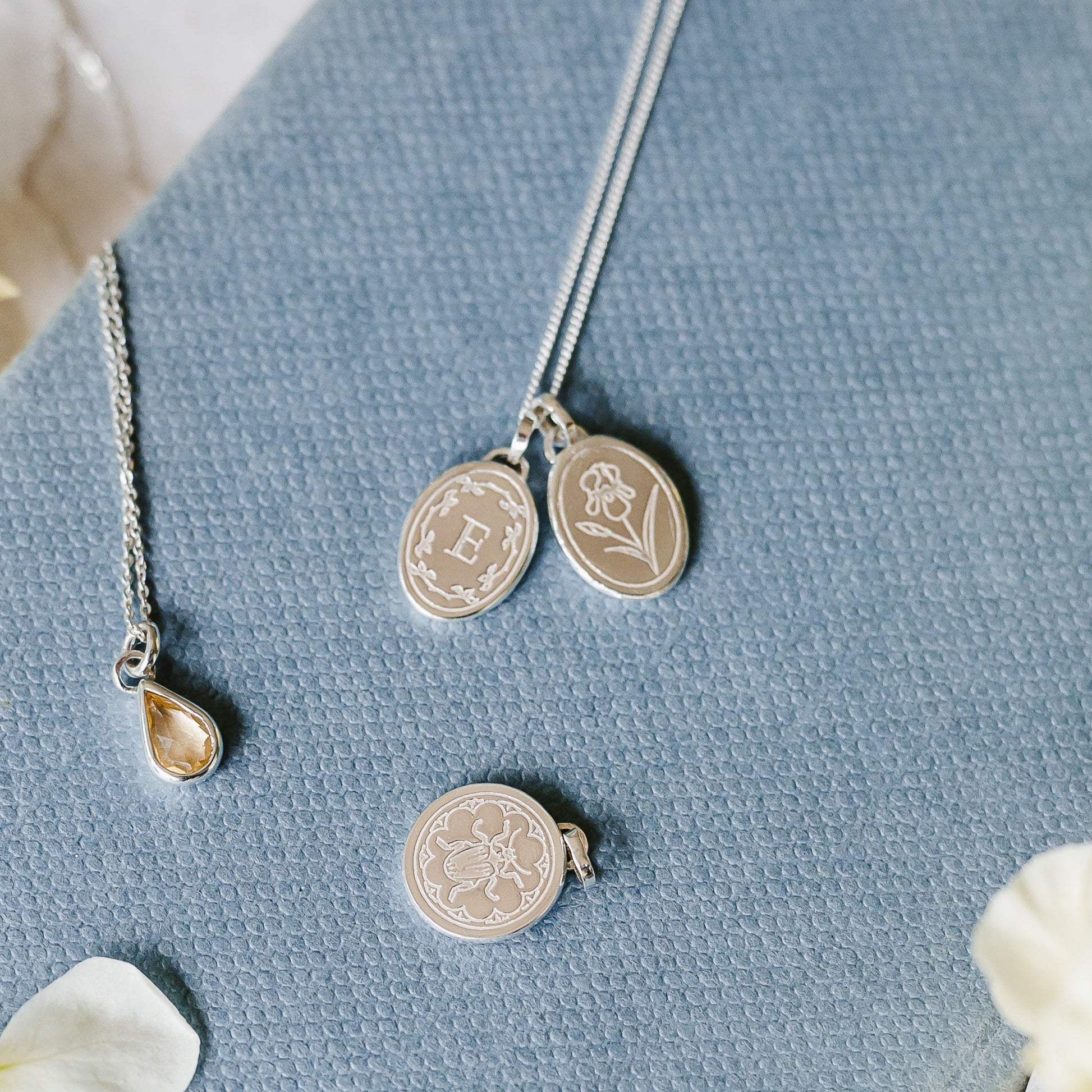 Sterling silver initial necklace stacked with Iris birth flower. A bee and a yellow teardrop charm also shown. All sterling silver pieces lying on a pale blue book cover.