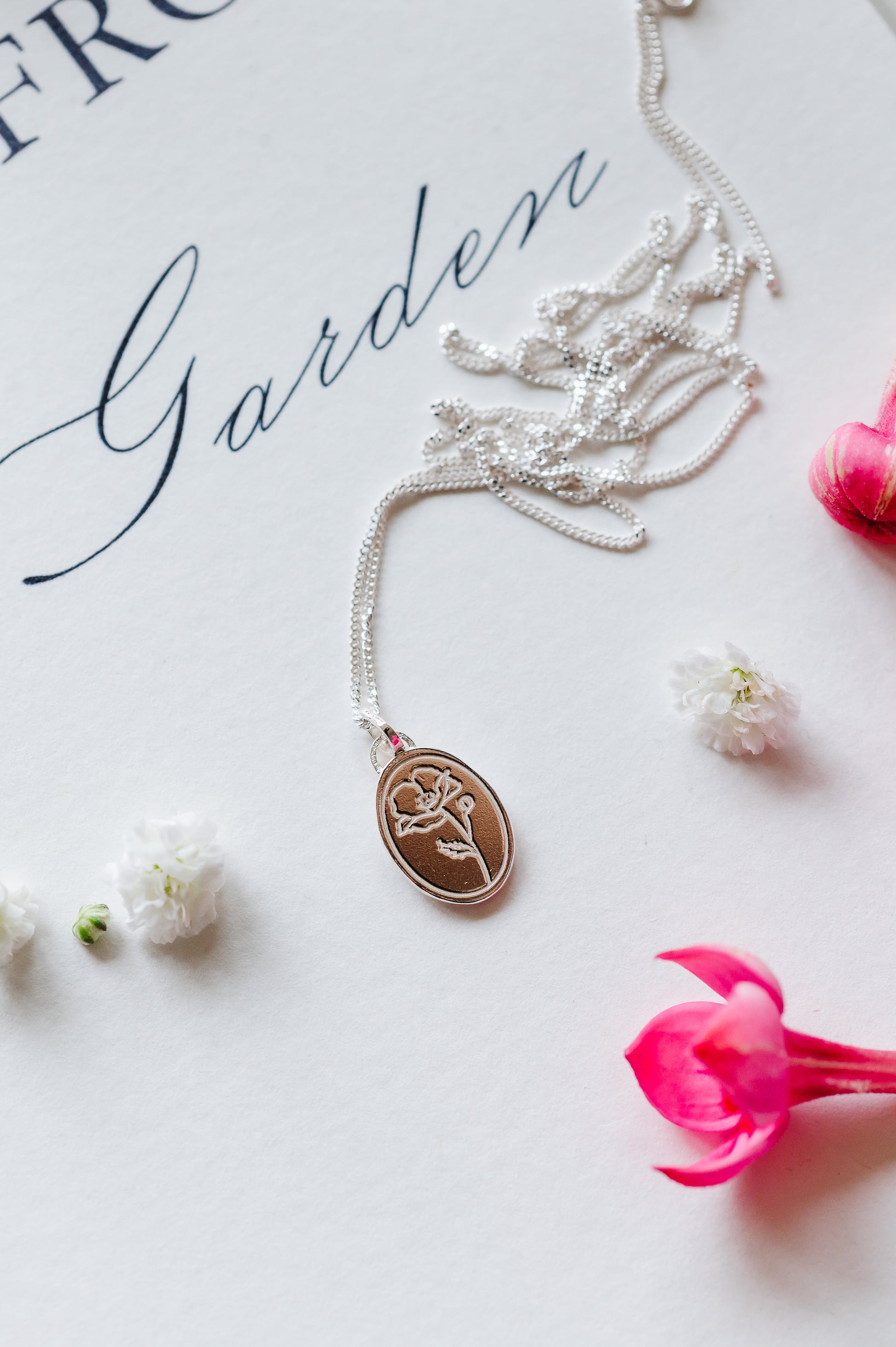 A sterling silver necklace with an oval coin pendant featuring a poppy design, displayed on a white surface with a chain and additional decorative flowers.