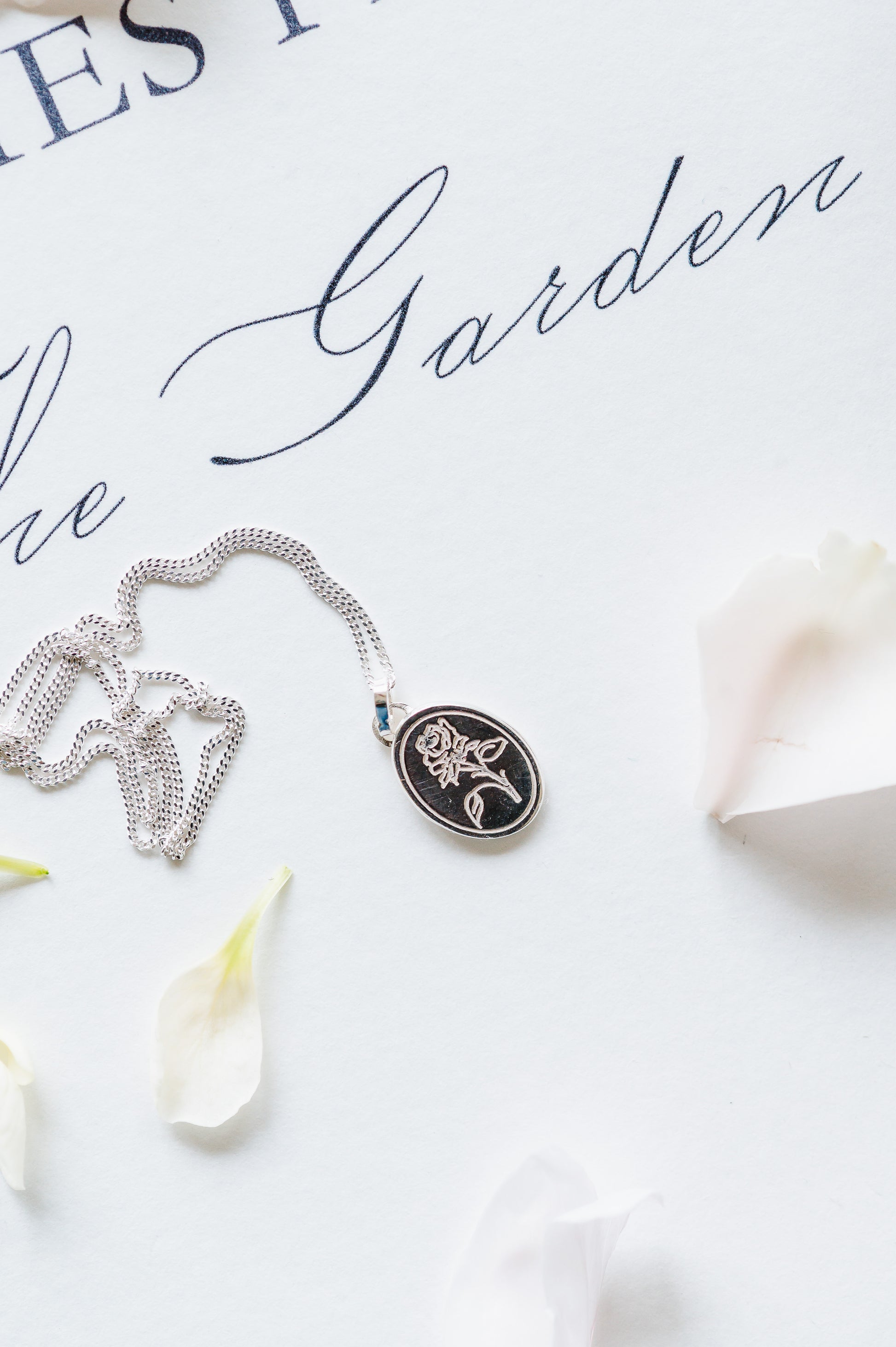 A sterling silver necklace with an oval coin pendant featuring a rose design, displayed on a white surface with a few petals around.