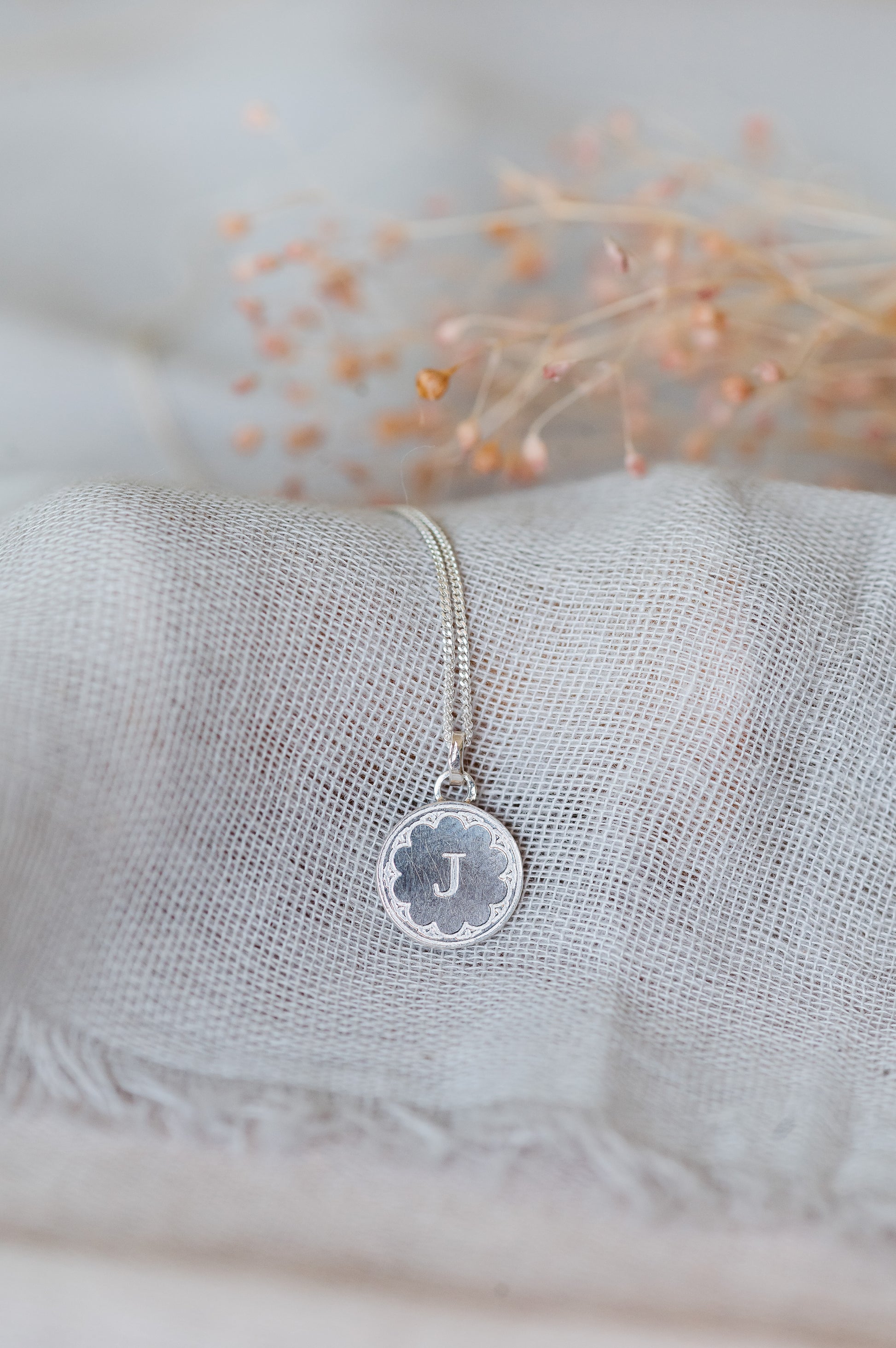 A round initial necklace in sterling silver, sitting on very pale blue linen with flowers in the background.