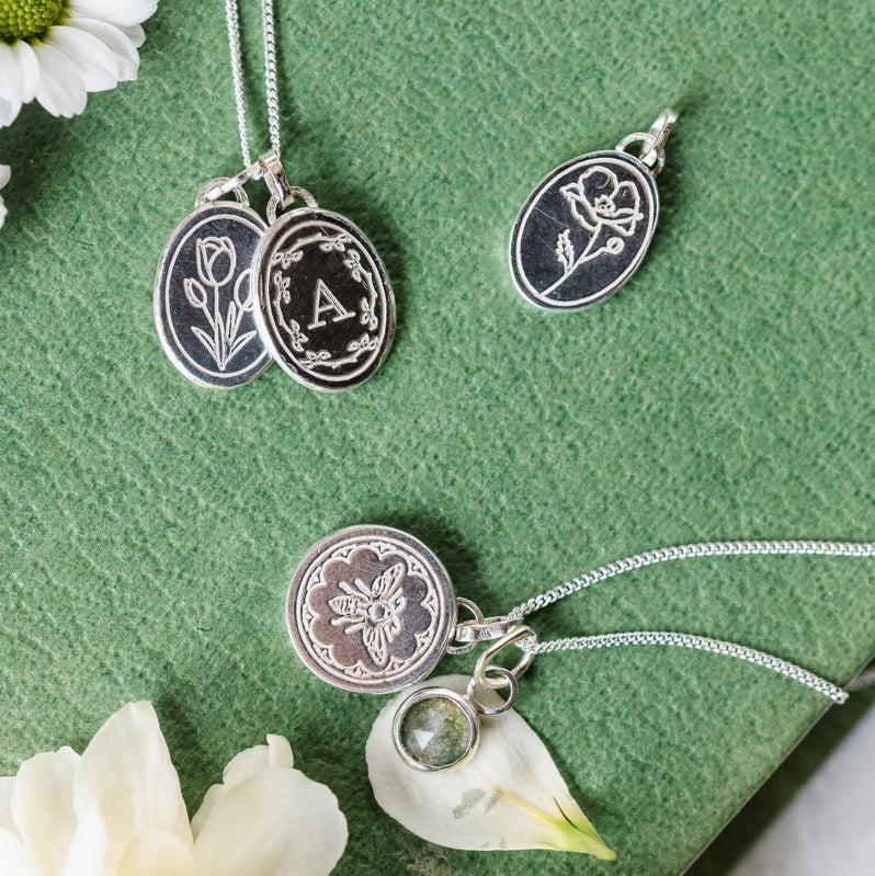 A selection of engraved necklaces, initial necklace, birth flowers and bee necklace sitting on a book cover with flowers.