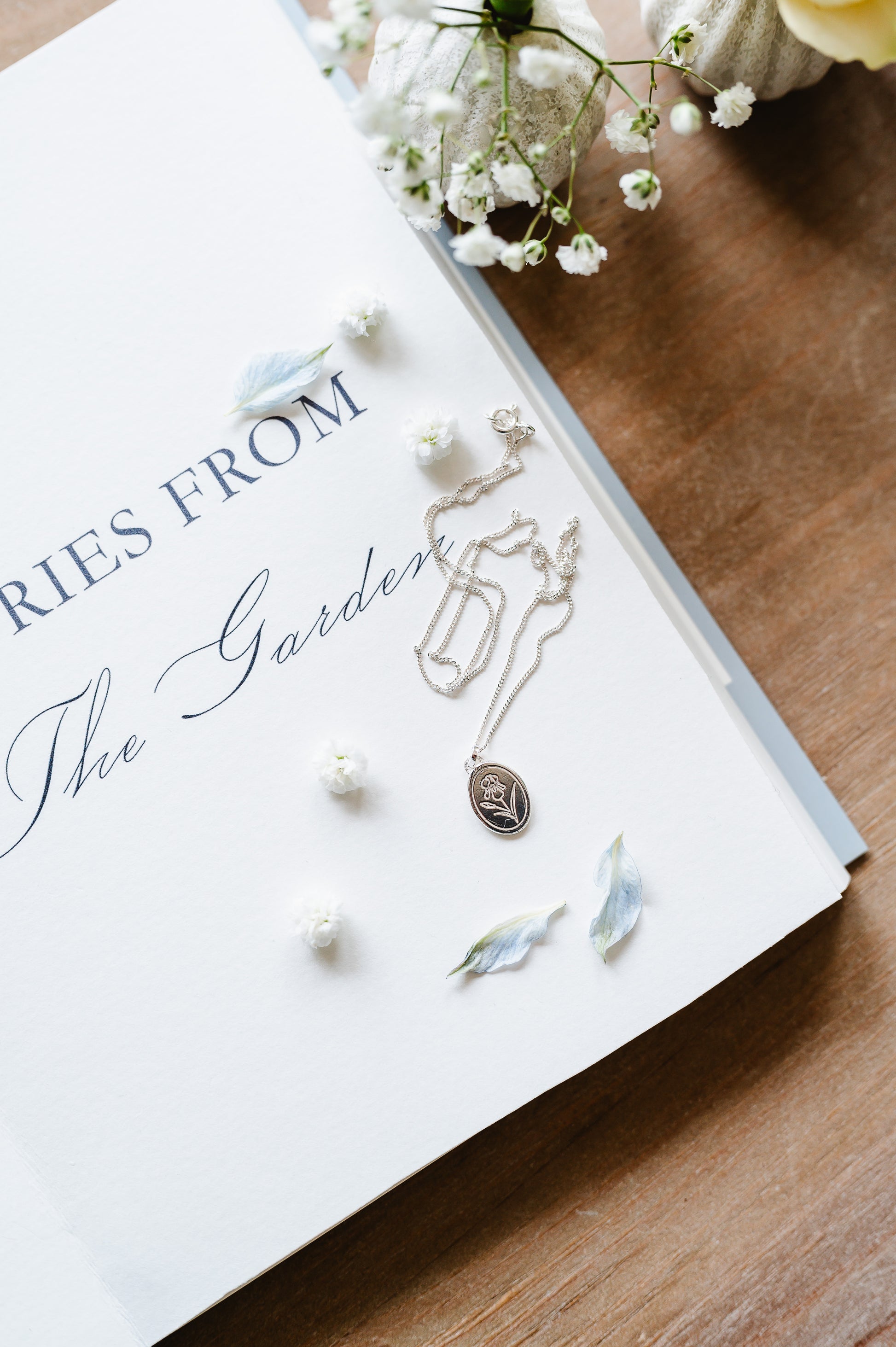 A dainty necklace with an iris flower in sterling silver, lying on a book with flowers in the background. 