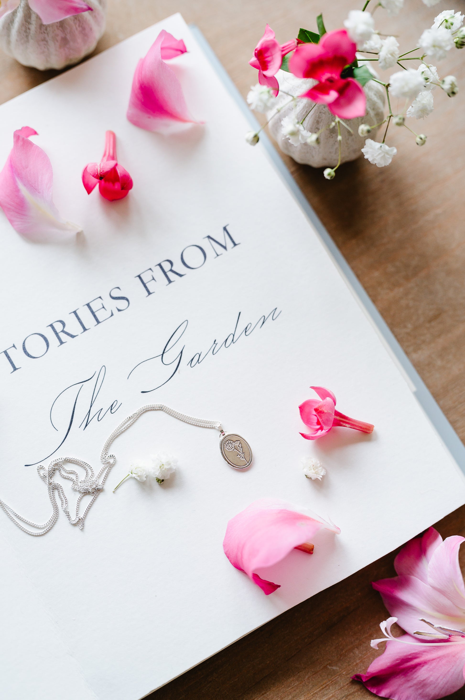 A dainty sterling silver necklace with a cosmos flower lying on a book. Flowers in the background. 