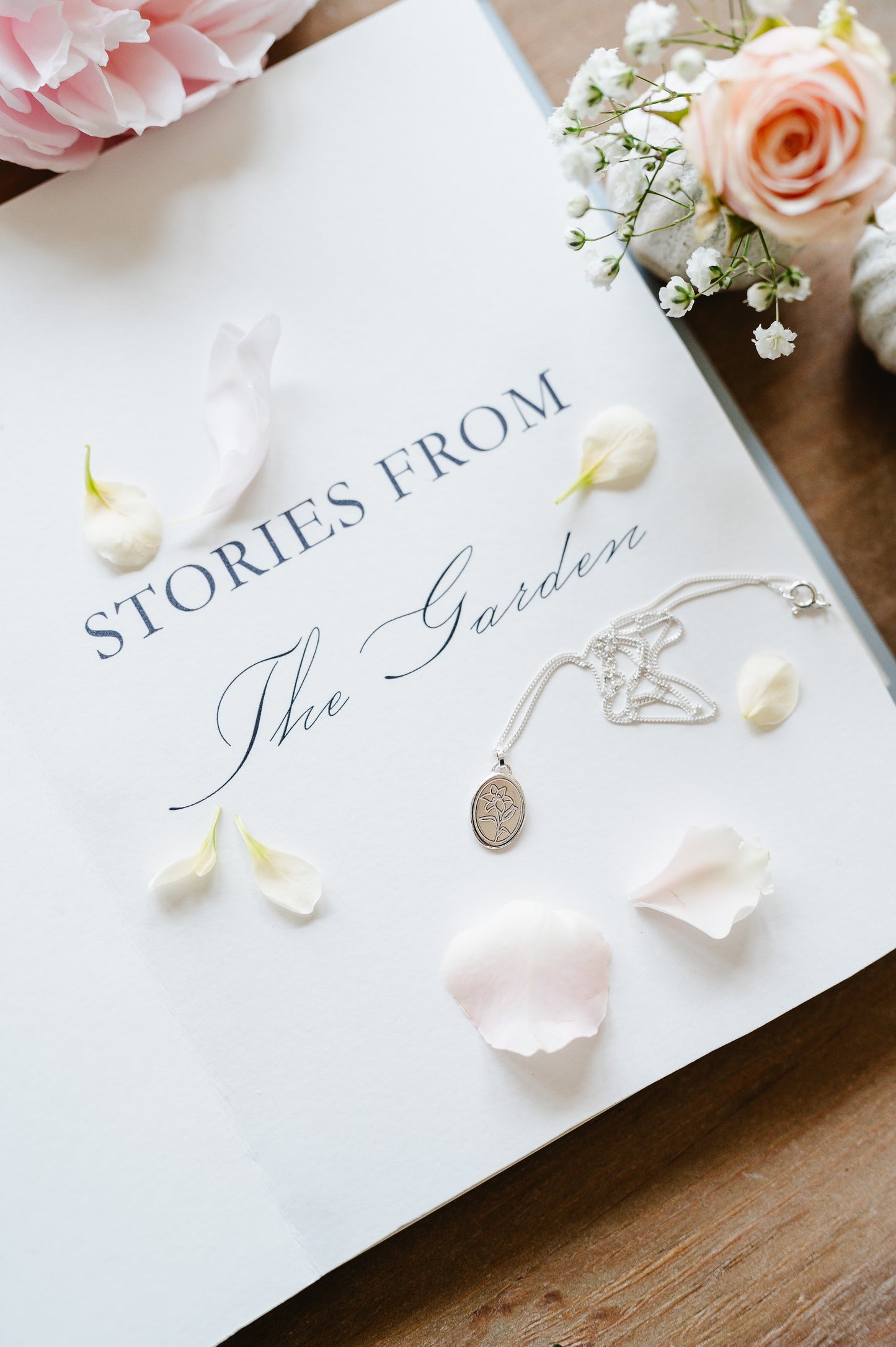 A dainty necklace in sterling silver with a lily, lying on an open book. Petals are strewn across the book.