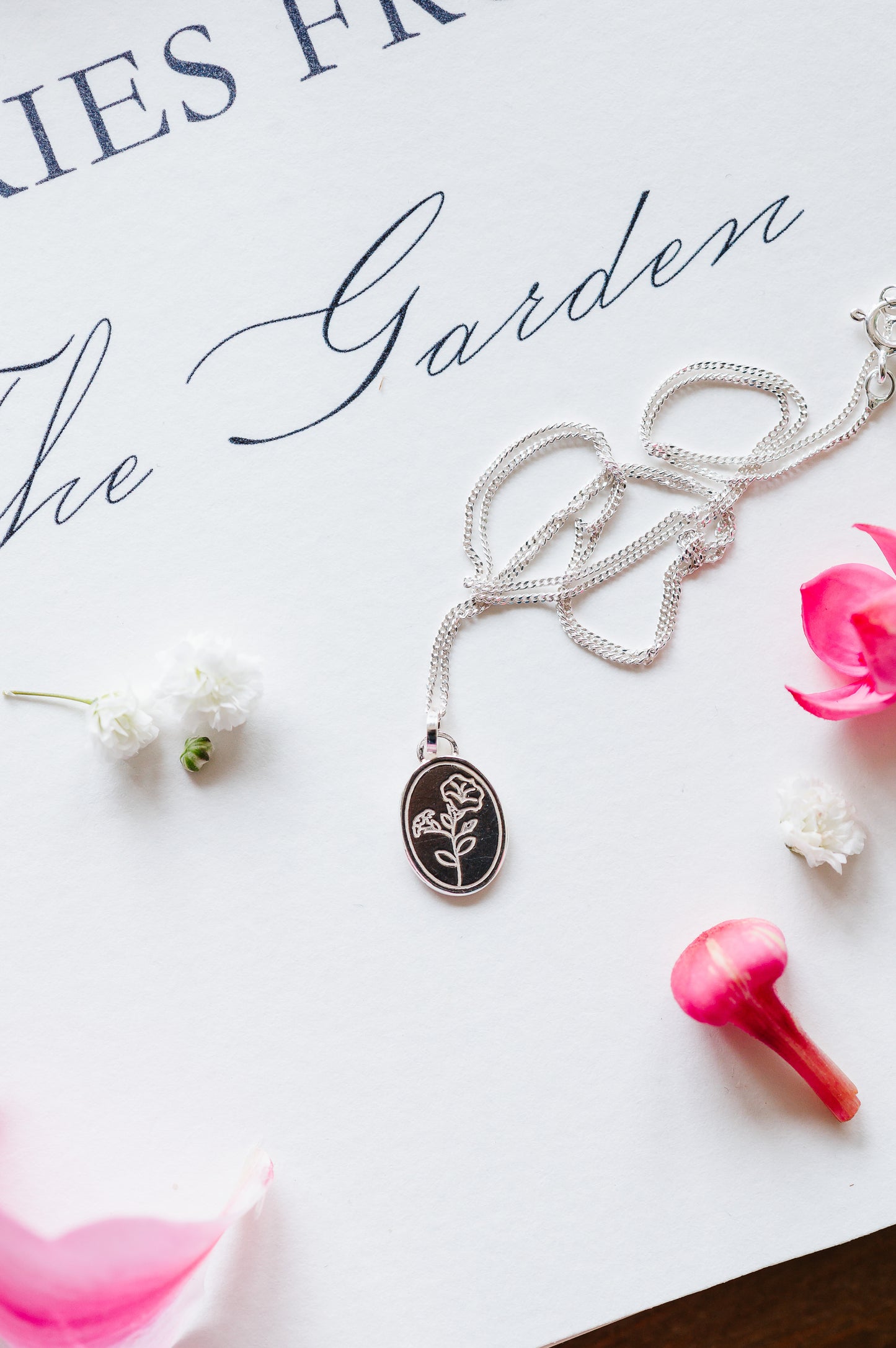 A flower necklace with a morning glory in sterling silver, lying on a white background with some scattered petals. 