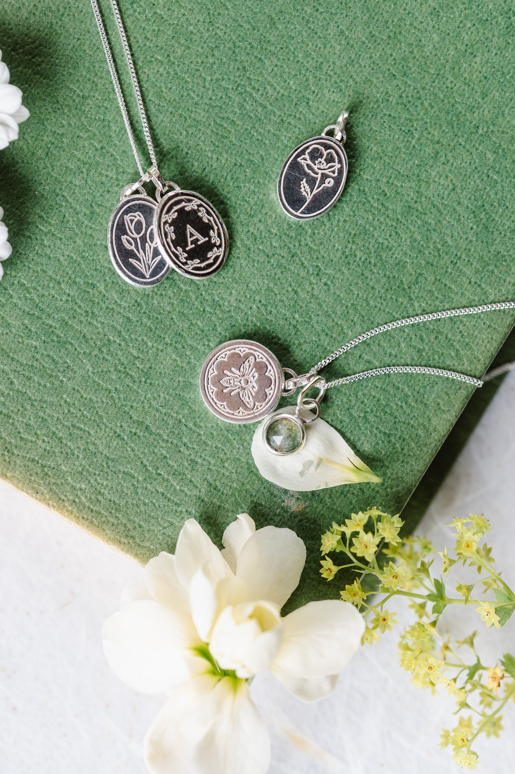 A sterling silver initial necklace stacked with a birth flower, lying on a book with some other sterling silver necklaces.