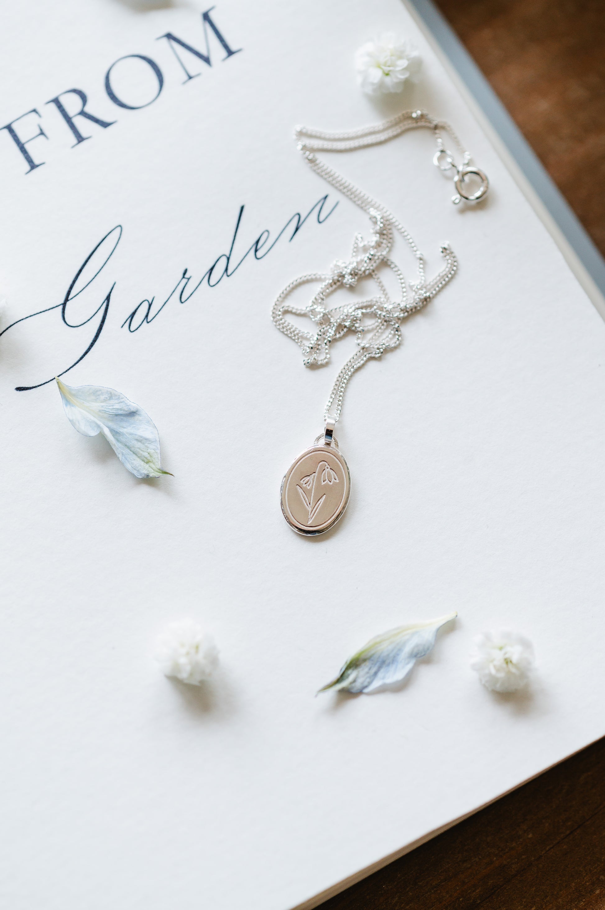 A sterling silver necklace with a snowdrop flower on a book page, surrounded by petals