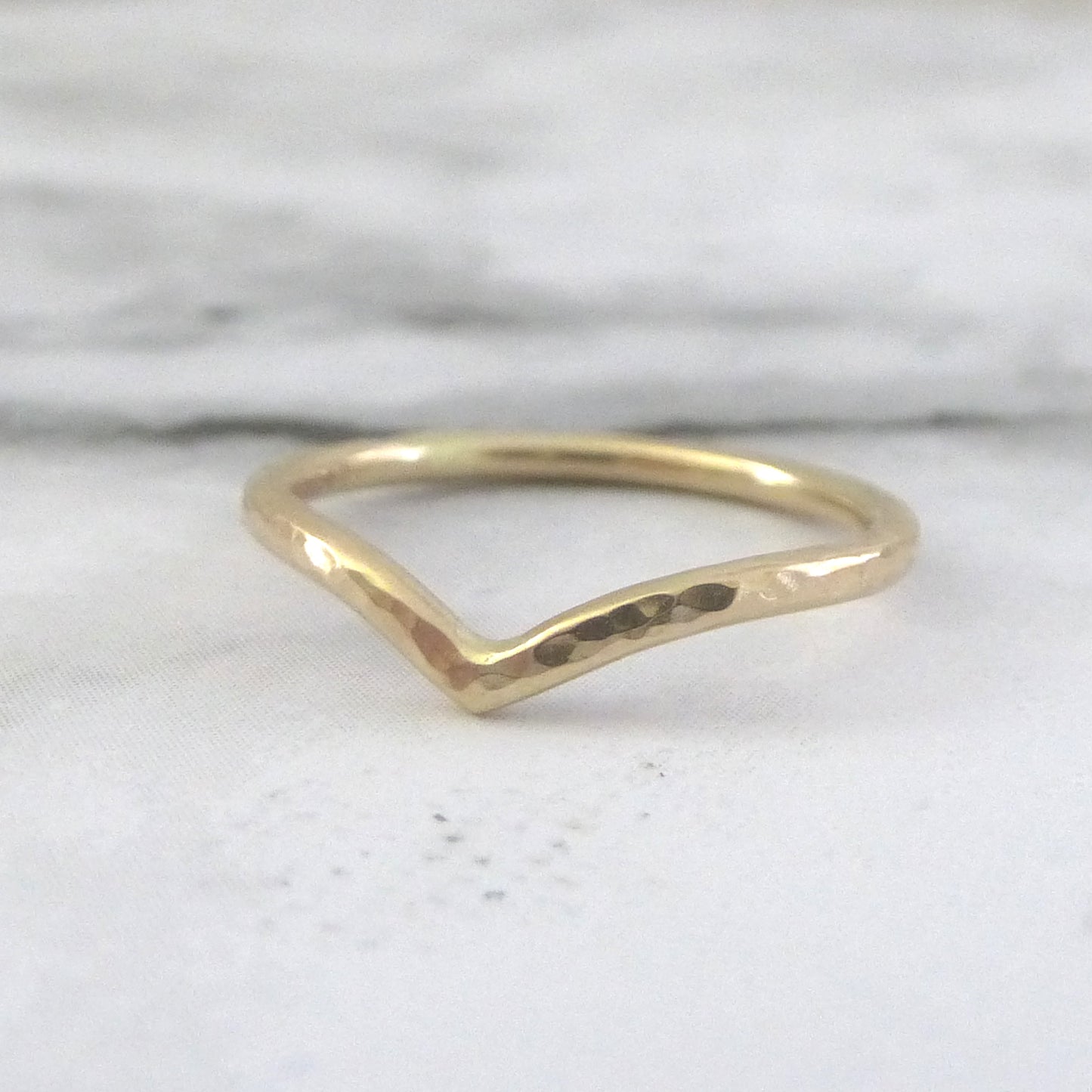 A subtle yellow gold wishbone ring with a hammered texture, displayed against a marble paper background.